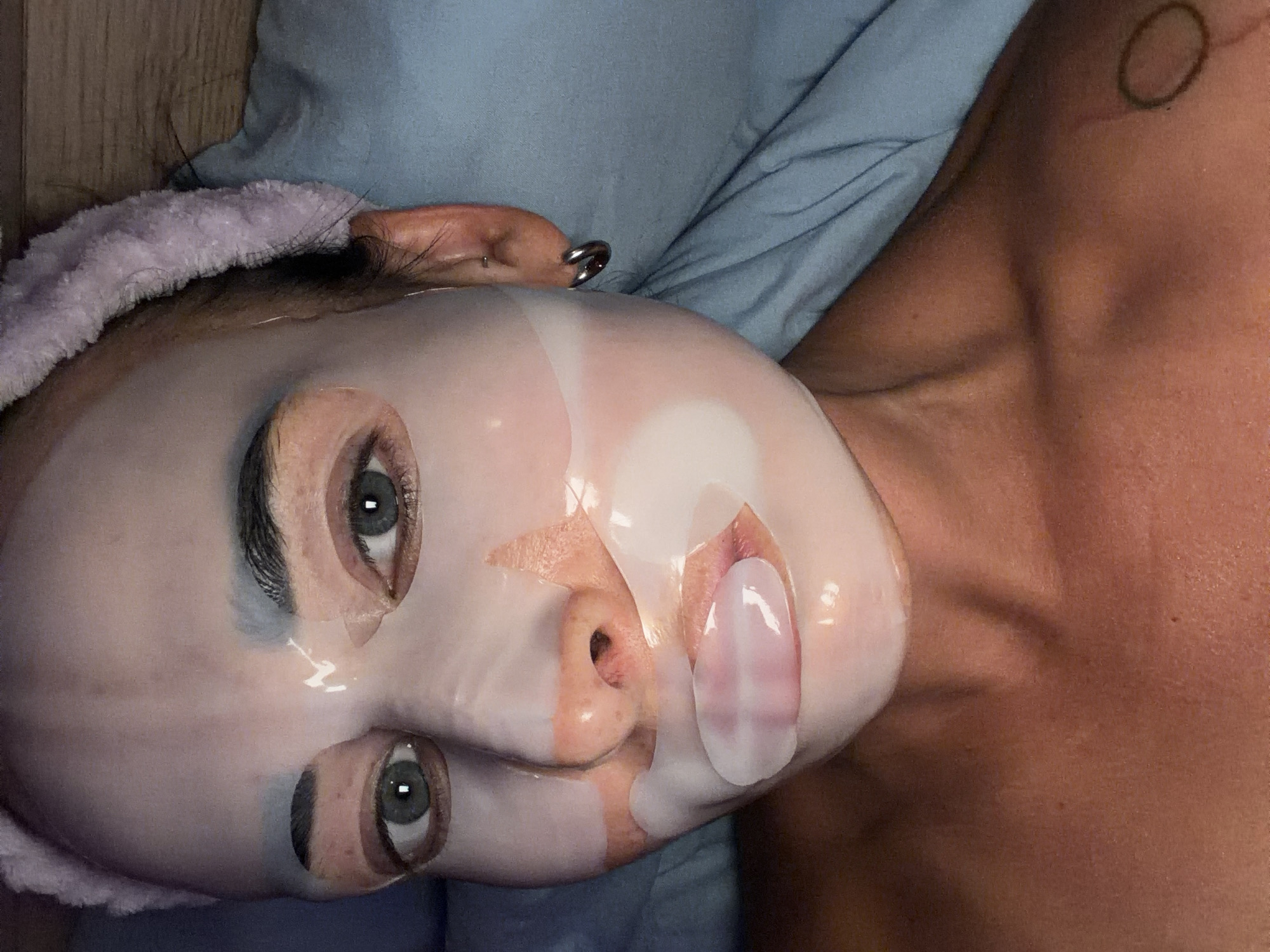 A person lies on a blue surface, possibly a bed or cushion. They have a sheet mask on their face and are wearing a soft pink headband. The mask covers most of their face except for the eyes, nose, and mouth. An earring is visible in their ear. A circular tattoo is on their upper chest/shoulder area.
There is no verifiable connection to the city of Saint Petersburg, Russia.