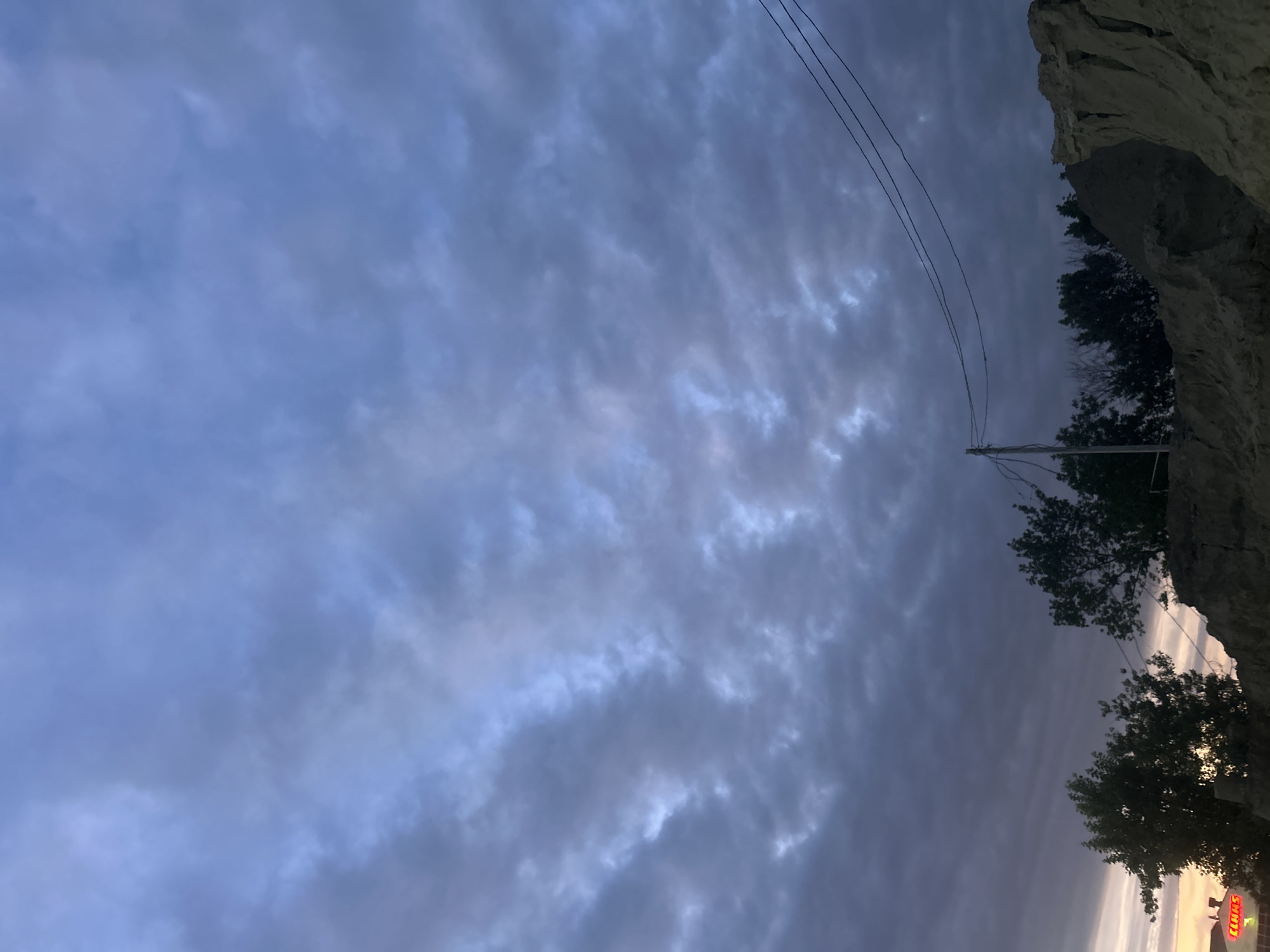 The image depicts a landscape at dusk in Krasnodar, Russia. The sky is filled with a mix of dark grey and lighter blue clouds, suggesting an overcast or stormy weather condition. In the foreground, there are several silhouetted trees and a rugged, earthy embankment. A utility pole with wires stretches across the frame, dividing the sky and connecting to the embankment. To the left, a portion of a building with illuminated signage, possibly a business, is visible. The overall scene conveys a somewhat somber and natural atmosphere characteristic of the outskirts of a city during twilight.