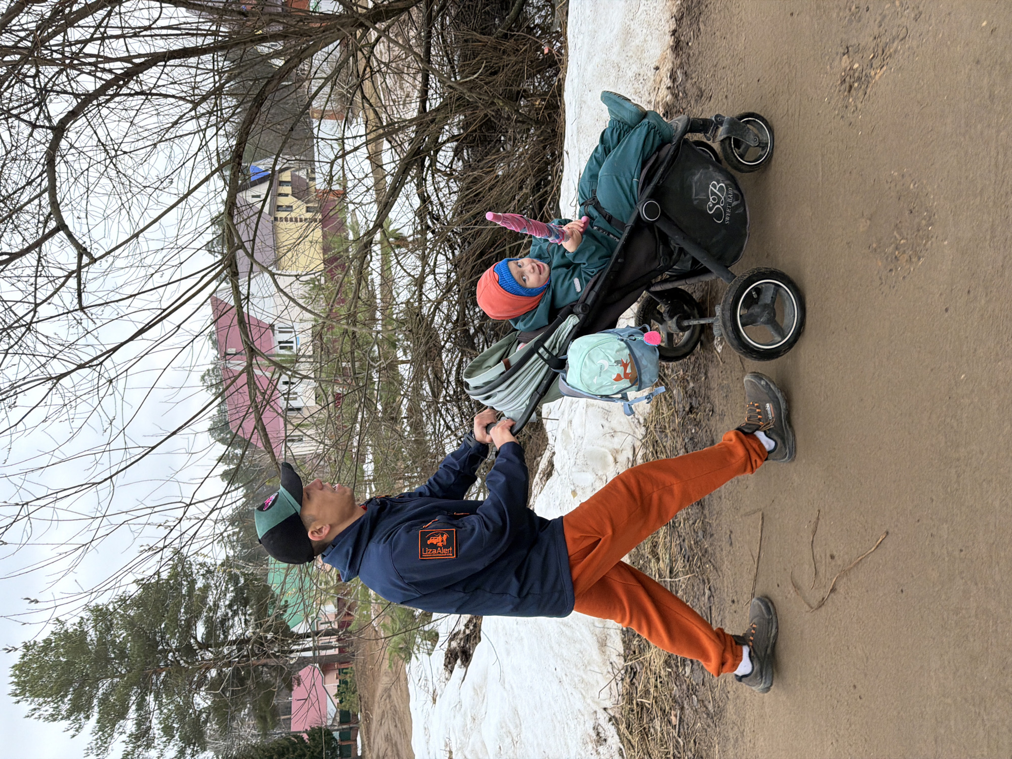 A man is pushing a baby stroller on a path in a park. The stroller contains a child wearing a red hat and green outfit, who is holding a pink object. The man is wearing a blue jacket, orange pants, a hat, and shoes. He is gripping the stroller handle with both hands and facing forward.
The path is dirt and there is snow on either side. Bare trees and branches are visible in the background, along with houses in the distance. The location appears to be a park near houses. There is a "Liza Alert" patch on the man's jacket.
The location is the city of Chaikovsky, Russia.