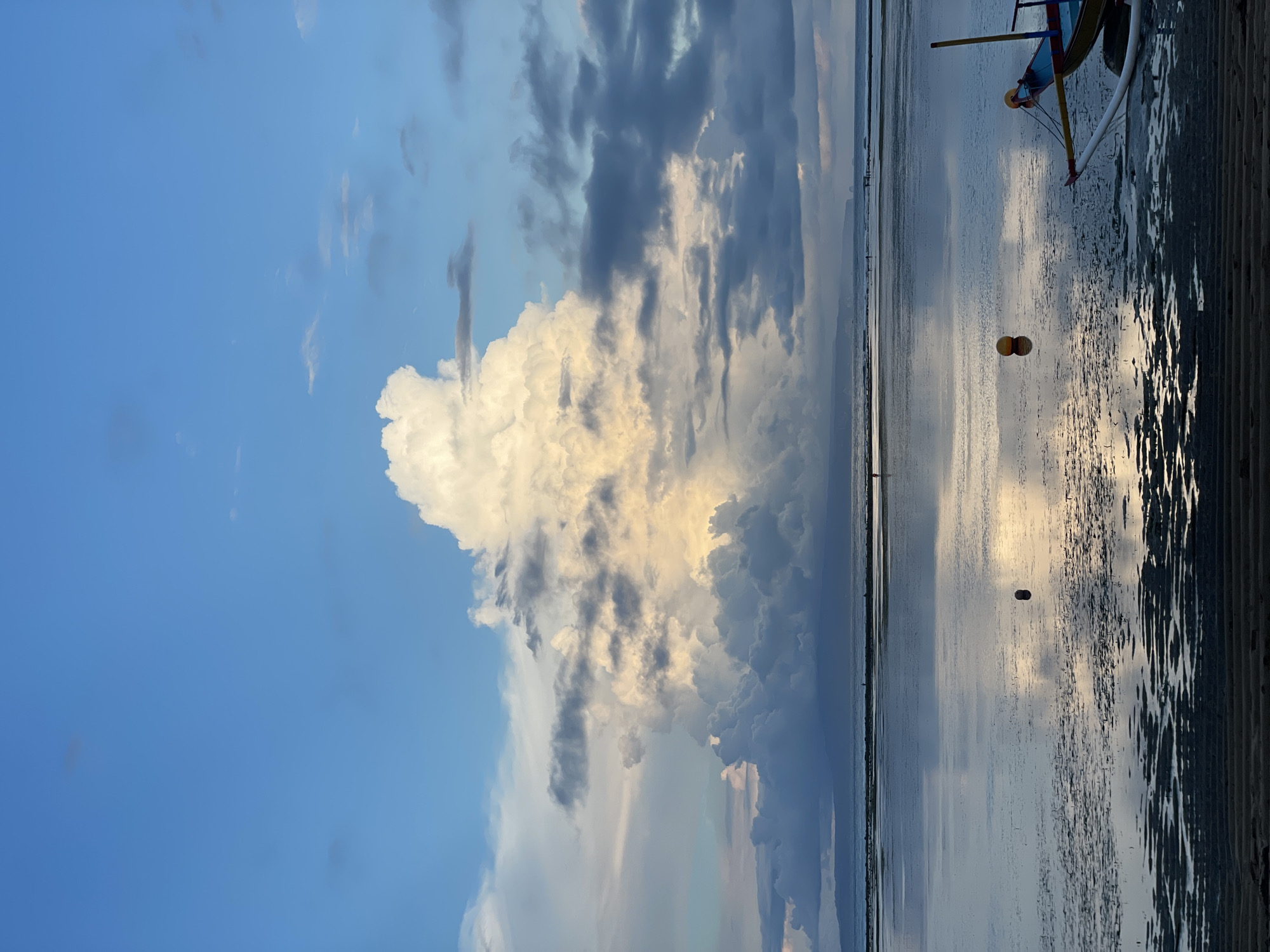 A sideways view shows a serene coastal scene likely near Denpasar, Indonesia, as it features elements typical of the region. A large, billowy cloud formation dominates the sky, juxtaposed against patches of blue. The water appears calm and shallow, reflecting the sky and clouds. A small outrigger boat rests near the edge of the water, with distinctive buoys attached. The foreground consists of dark, textured sand.