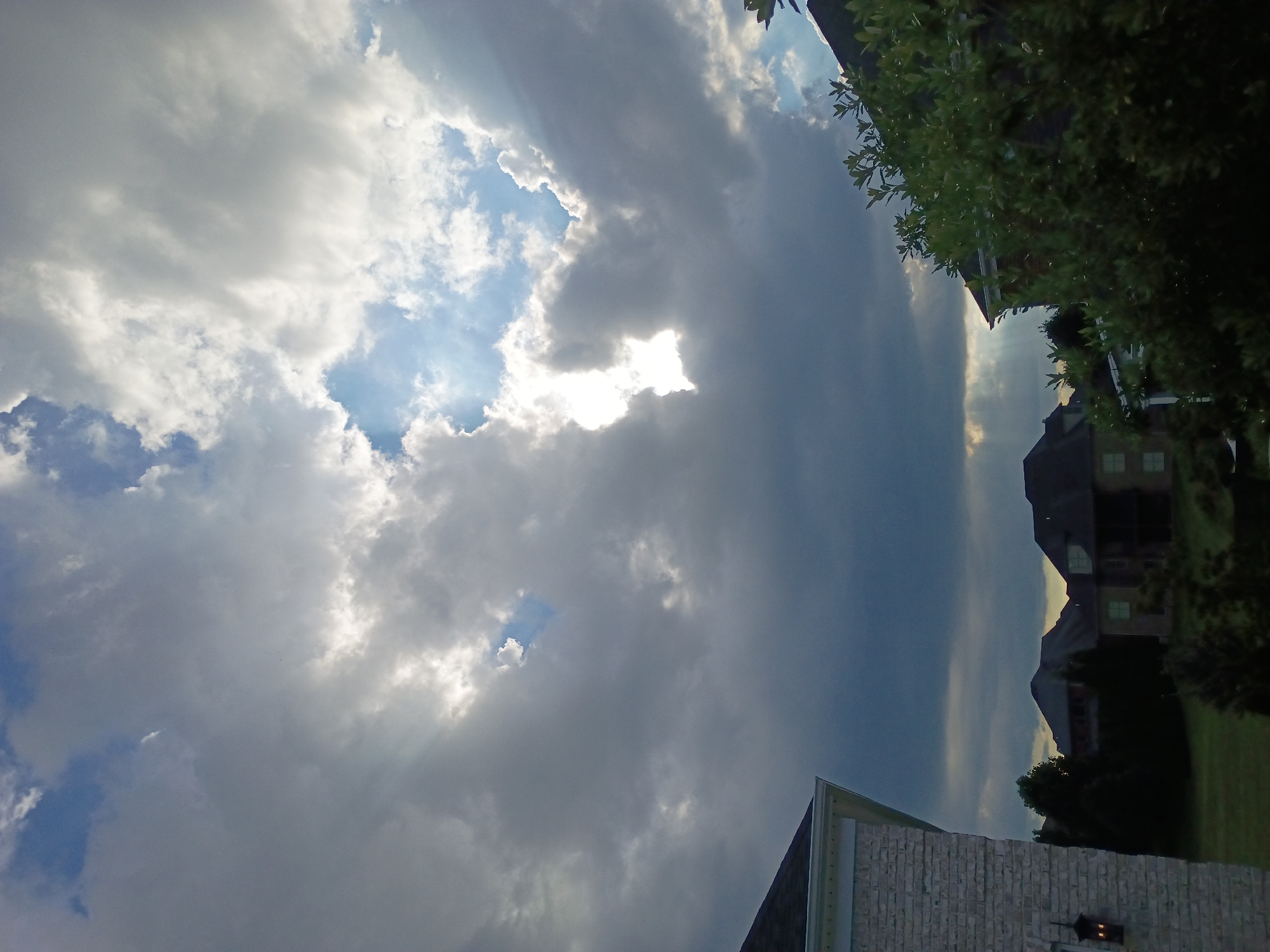The image captures a dramatic sky over a suburban residential area. The upper portion of the frame is dominated by a vast expanse of clouds, with a bright opening in the center revealing a patch of blue sky and sunlight streaming through. Shafts of light emanate from this opening, illuminating the surrounding clouds. Below the dramatic sky, the roofs and upper portions of several large houses are visible, nestled amongst dark green trees. In the foreground to the left, the corner of a light-colored brick house with a black lantern is partially visible. The overall scene suggests an impending storm or the aftermath of one, with the interplay of light and dark clouds creating a striking visual effect. The location is specified as Lexington, United States of America.
