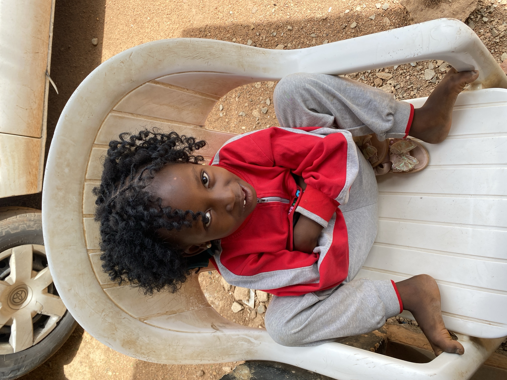 A young girl is sitting in a white plastic chair outdoors. She has dark curly hair and is wearing a red and gray tracksuit. Her feet are bare and appear dirty. The chair is slightly dirty, and the background is a dirt surface. The front of a car is partially visible on the left side of the frame. The location is presumably Kuru Karama, Nigeria.