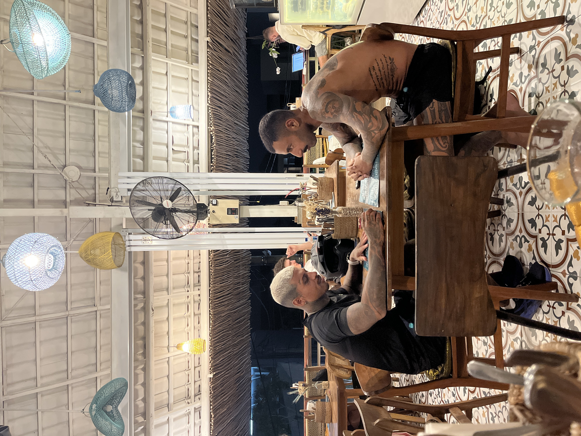 Two men are seated at a table in an outdoor restaurant in North Kuta, Indonesia. The man on the left, with bleached blonde hair and extensive tattoos, is wearing a black t-shirt and looking down at a menu on the table. The man on the right, with dark hair and a mustache, is shirtless and has tattoos covering his chest and arms. He is looking towards the left, with his hands clasped on the table. The restaurant has a tiled floor with a patterned design, wooden tables and chairs, and a ceiling covered with woven palm leaves. Several decorative hanging lights and a ceiling fan are visible. In the background, other diners and restaurant staff can be seen. The overall atmosphere suggests a casual dining experience.