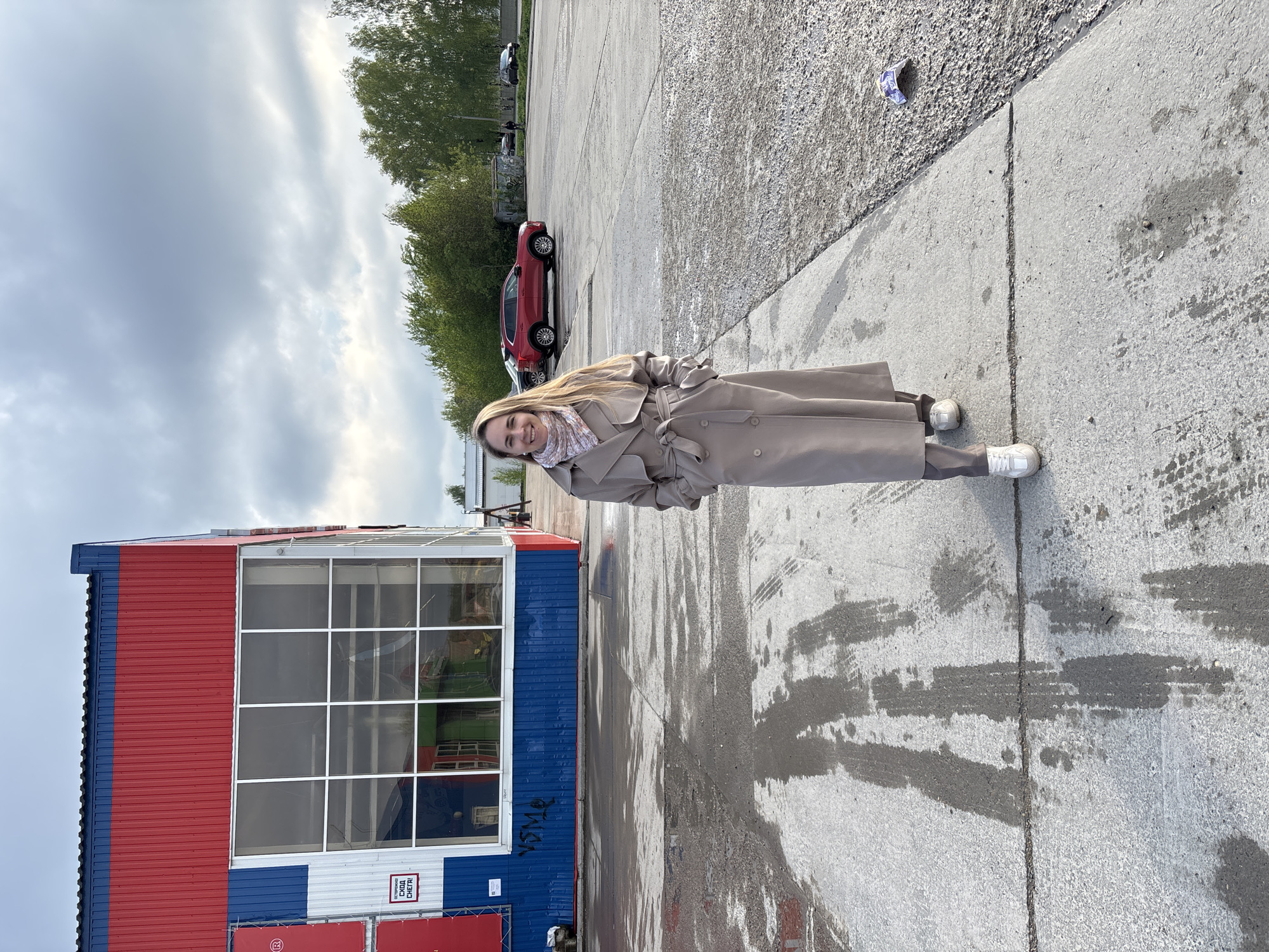 A woman stands in a parking lot in front of a blue and red building. The building is located in the city of Chaikovsky, Russia. A red car is parked in the background. The woman is wearing a beige coat and white sneakers. She is smiling and looking at the camera. The ground is concrete and wet. The sky is cloudy.