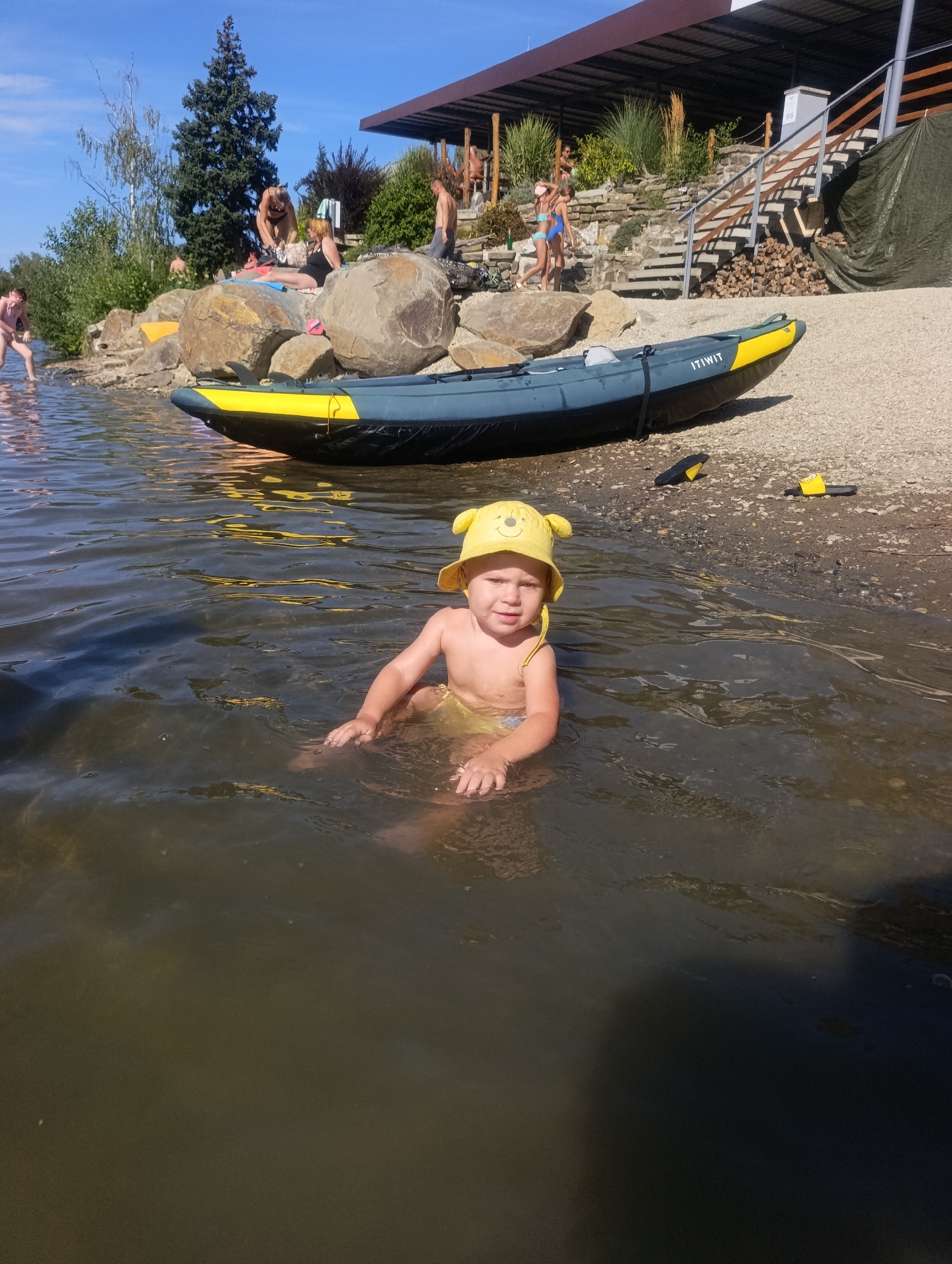 A young child sits in shallow, murky water at the edge of a lake or river, wearing a yellow sun hat with bear ears. The child is partially submerged and appears to be playing in the water. In the background, a blue and yellow kayak rests on a pebbly shore. Beyond the shore, people are visible, some sitting on large rocks and others walking near a building with a covered outdoor area and a set of stairs leading up a grassy embankment. The scene suggests a warm, sunny day, likely during the summer, with people enjoying outdoor recreation. The sky is clear and blue. The child's expression is neutral, but the overall atmosphere of the image is relaxed and familial. The word "ITIWIT" is visible on the side of the kayak. The location is Křelov, Czechia.