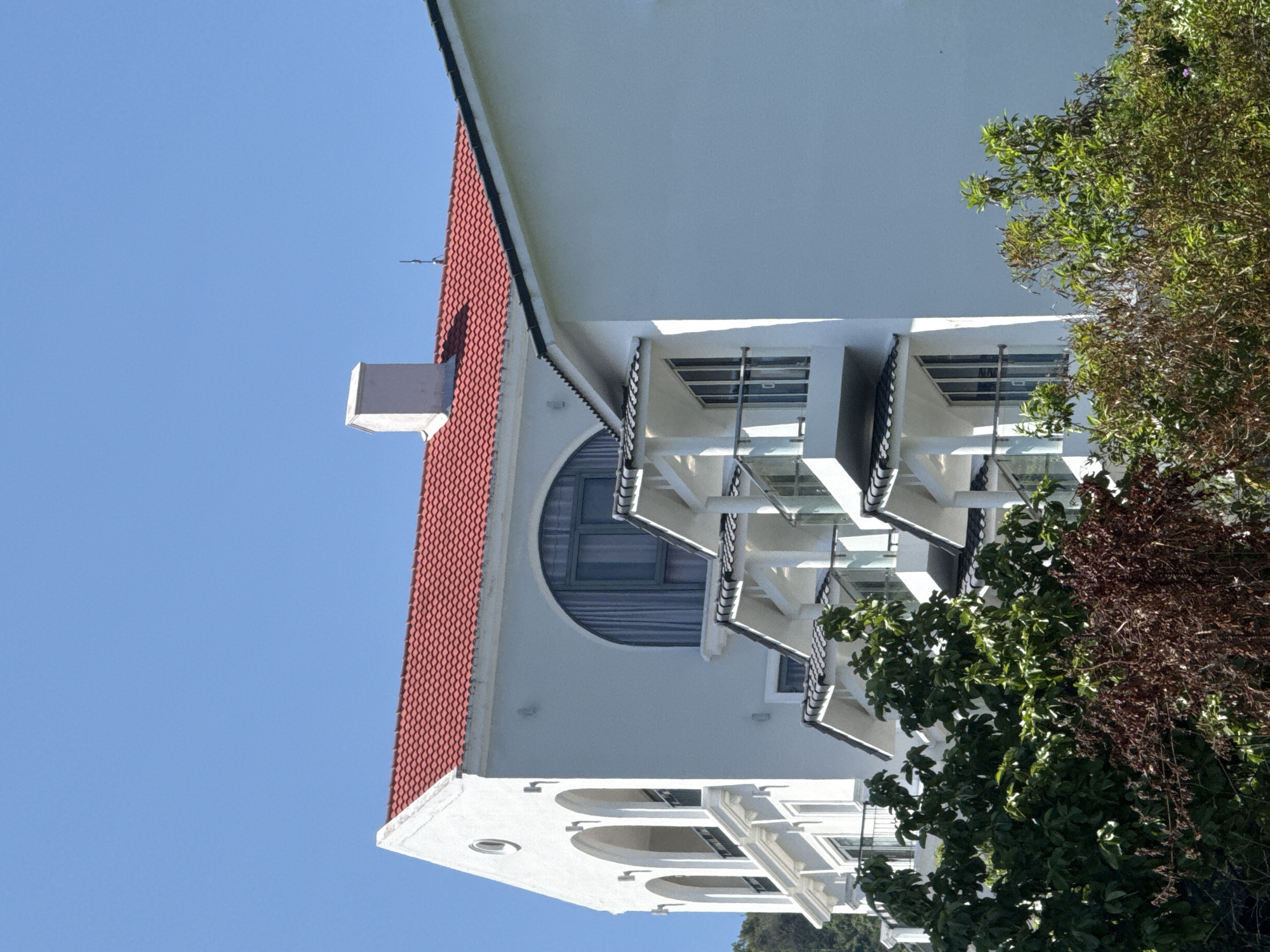 This is a low-angle, medium shot of a white building with a red tiled roof against a clear blue sky. The building has a distinctive architectural style, with arched windows on the upper left side, several balconies with glass railings, and tiered sections of rooflines. The building appears to be modern, possibly a hotel or residential complex. Green and brown foliage is visible in the foreground, partially obscuring the lower portion of the building. The overall scene suggests a bright, sunny day in a coastal or hilly area. The image does not contain any people or animals.