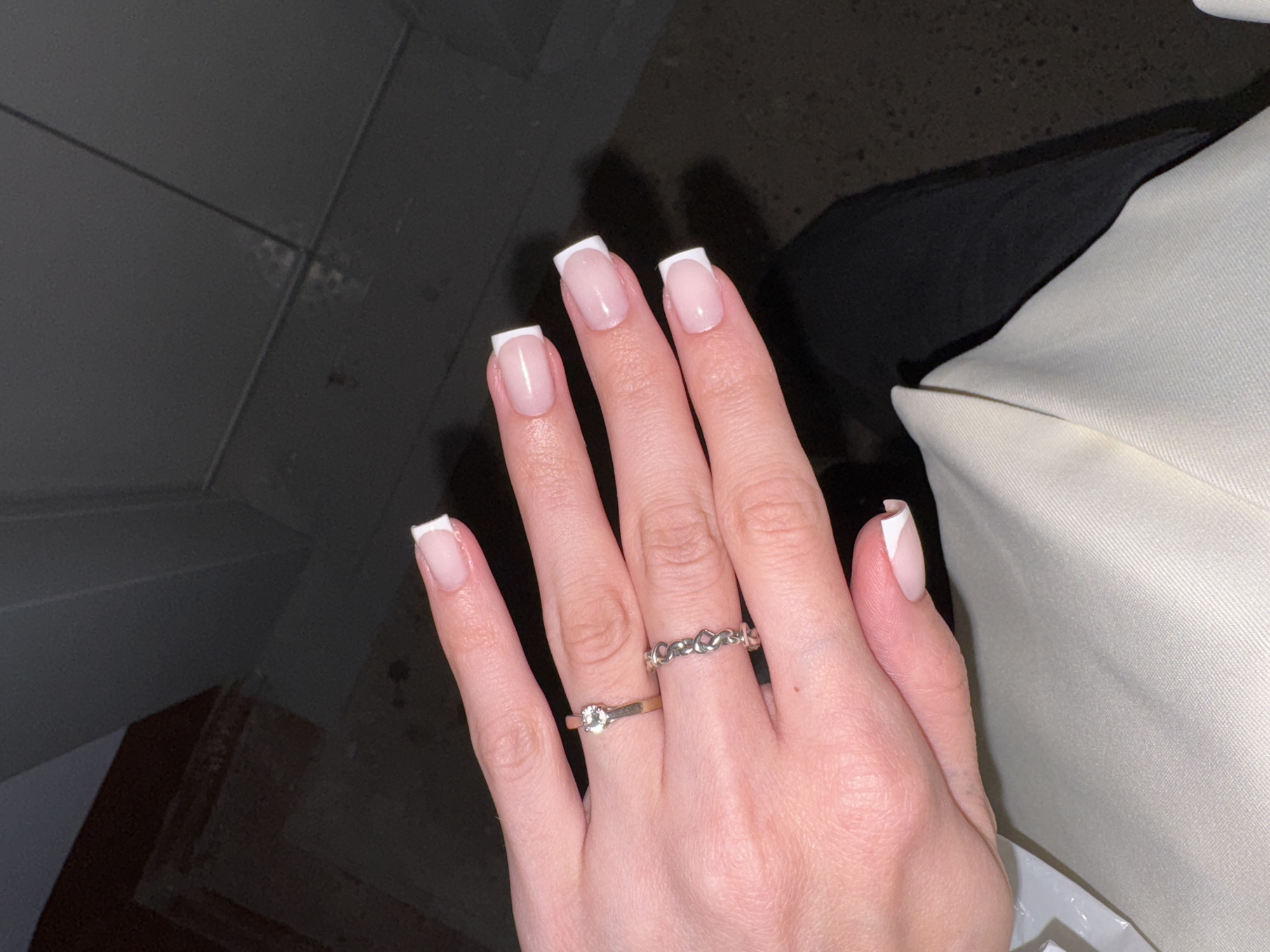 In an indoor setting, likely a hallway or staircase area, a close-up shot focuses on a person's hand adorned with a French manicure. The nails are sculpted into a square shape with a distinct white tip. Two rings are visible on the fingers: a thin gold band with a single clear stone on the ring finger, and a silver band with a linked heart design on the middle finger. The background is dimly lit and appears to consist of dark walls and a staircase. The lighting suggests it is evening or night. The location is identified as Perm, Russia, though no specific landmarks or context are provided within the image to confirm this.