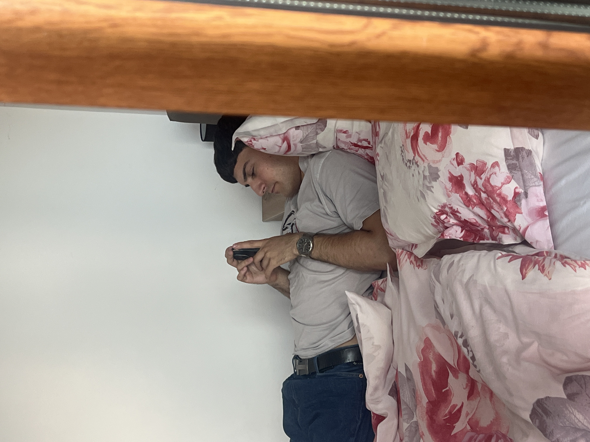 A man is lying in a bed, looking at a mobile phone in his hands. He is wearing a gray t-shirt, blue jeans, and a black belt with a silver buckle. He has a watch on his left wrist. The bed has a white sheet and a pillow with a floral pattern. The man is under a wooden beam. The location cannot be determined to be Varna, Bulgaria, from the visual information provided.