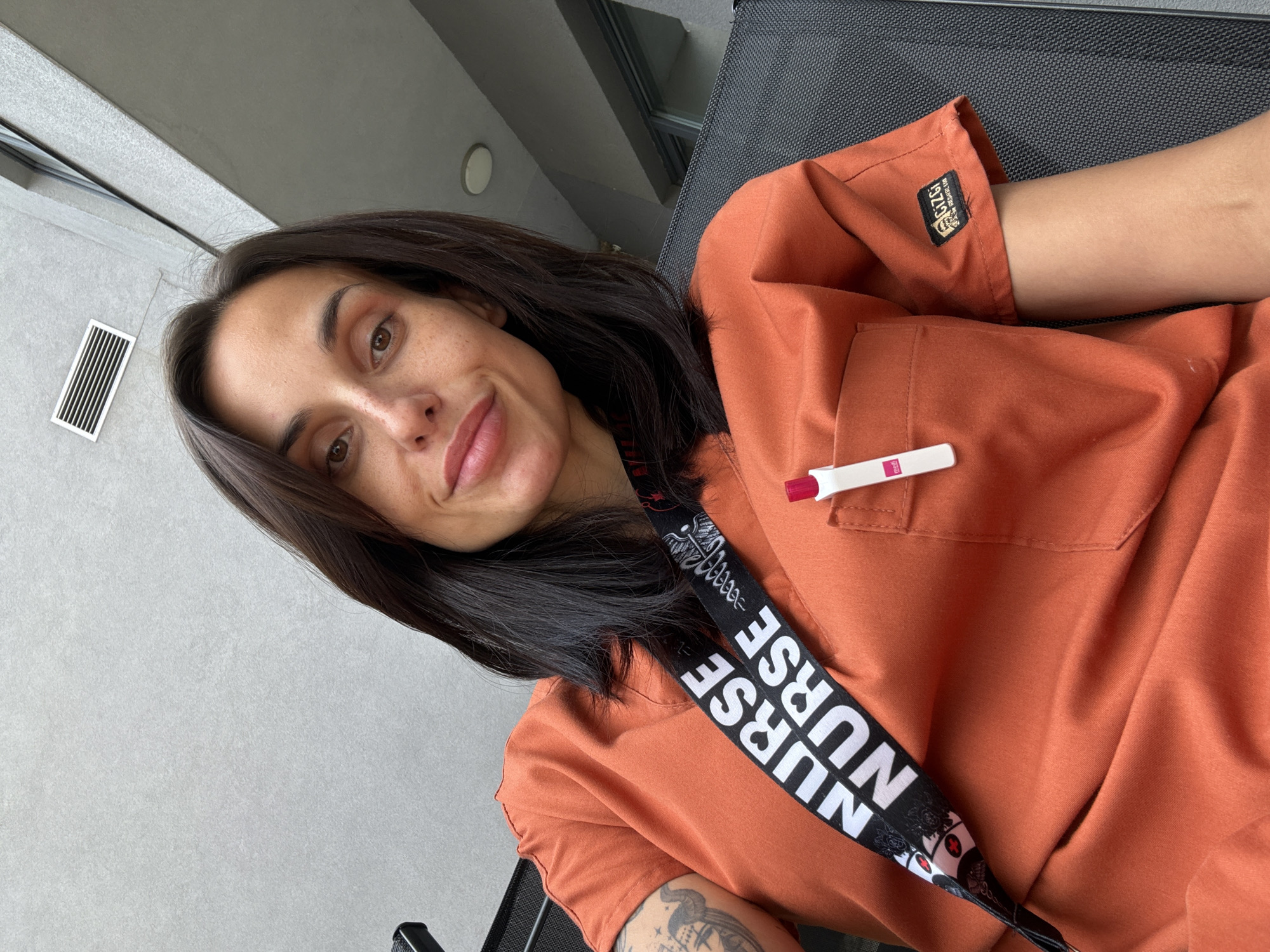 A woman with brown hair and eyes is wearing orange scrubs with a "Nurse" lanyard and a pregnancy test sticking out of the pocket. She is smiling slightly. The background appears to be a balcony or outdoor area with a gray wall and a window air conditioning unit. The location is unknown and there is no way to know where Pleven, Bulgaria is located.