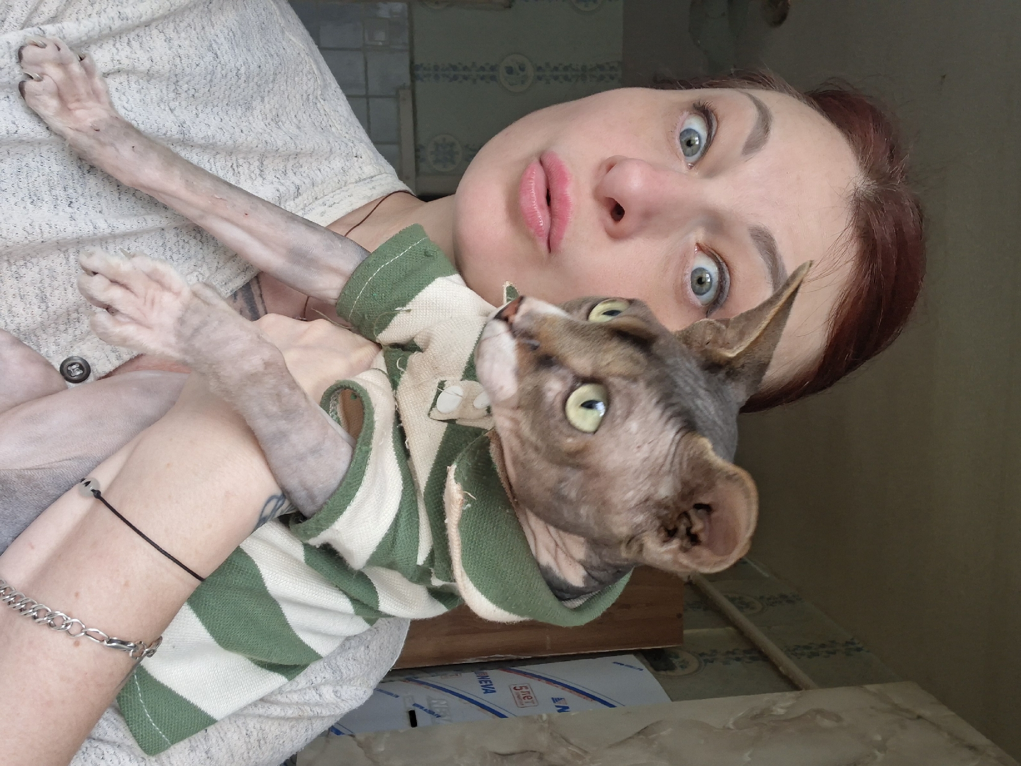 A medium shot captures a woman with auburn hair and wide, blue eyes, puckering her lips at the camera. She is holding a hairless cat, which is wearing a green and white striped shirt. The cat has large ears and green eyes. The woman is wearing a cream-colored, textured sweater. The background appears to be an indoor setting with a wall covered in light blue patterned wallpaper. To the right of the woman, a portion of a wooden cabinet and a white box with blue lettering are visible. The lighting suggests an overcast day or indoor lighting. The overall scene is candid and appears to be taken in a residential setting within Забалоцкі сельскі Савет, Belarus.