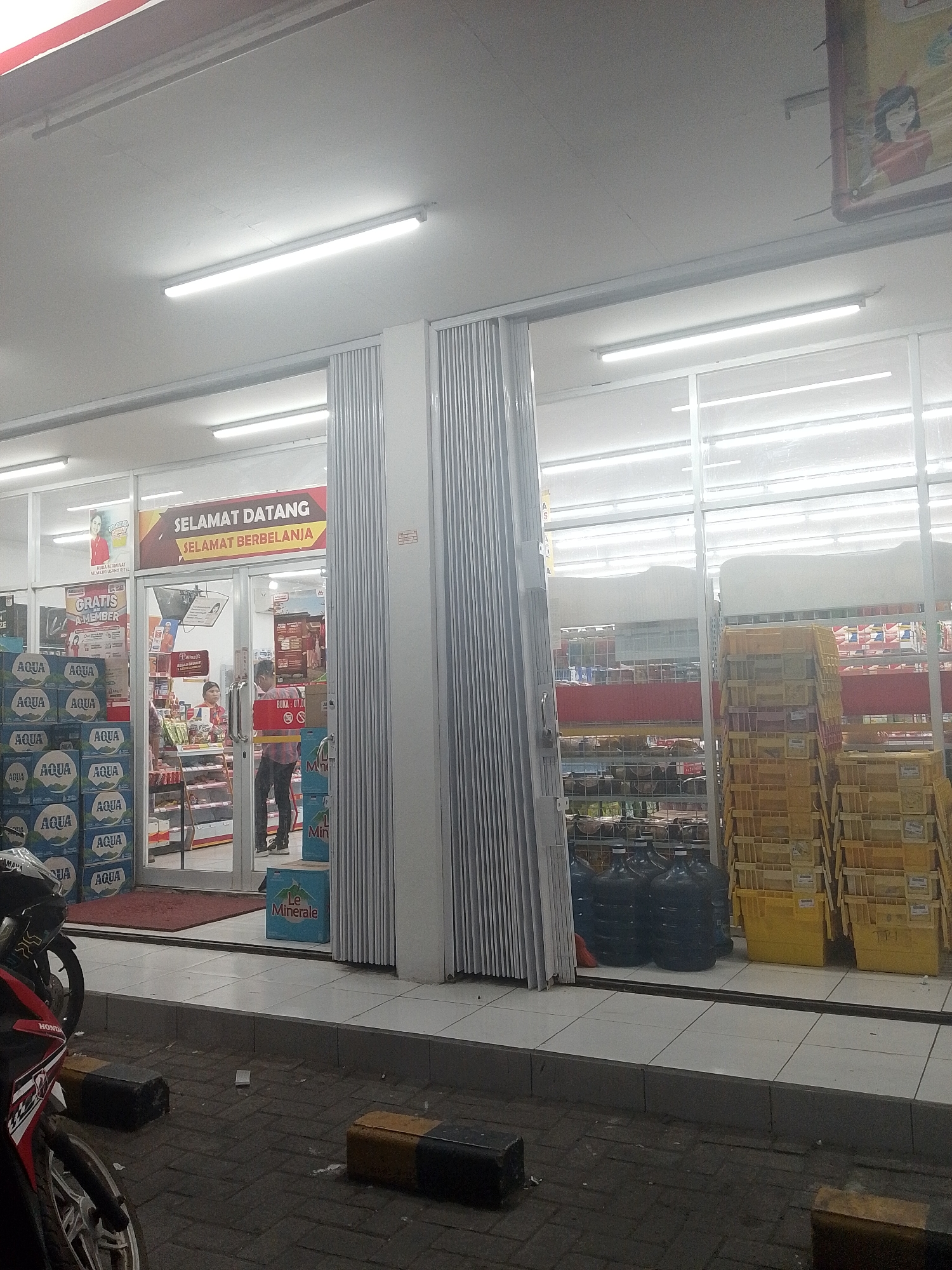 The image captures the exterior of a convenience store, likely located in Uwuran Satu, Indonesia, during what appears to be evening hours due to the artificial lighting. The store's facade features a large glass storefront with a retractable metal gate partially open. Above the entrance, a prominent banner reads "SELAMAT DATANG SELAMAT BERBELANJA" (Welcome, Happy Shopping).
Inside the store, visible through the glass, shelves are stocked with various products. A cashier, a woman wearing a red and white uniform, stands behind a counter. A male customer, wearing a plaid shirt and dark pants, is interacting with the cashier, presumably making a purchase. The interior lighting is bright and fluorescent.
To the left of the entrance, stacks of bottled water with "AQUA" branding are visible. To the right of the entrance, several large, dark blue water jugs are present, along with yellow crates stacked high. Outside the store, on the pavement, a motorcycle is parked. The ground is paved with tiles and dark asphalt. The overall scene suggests a typical retail environment in an Indonesian urban or suburban setting.