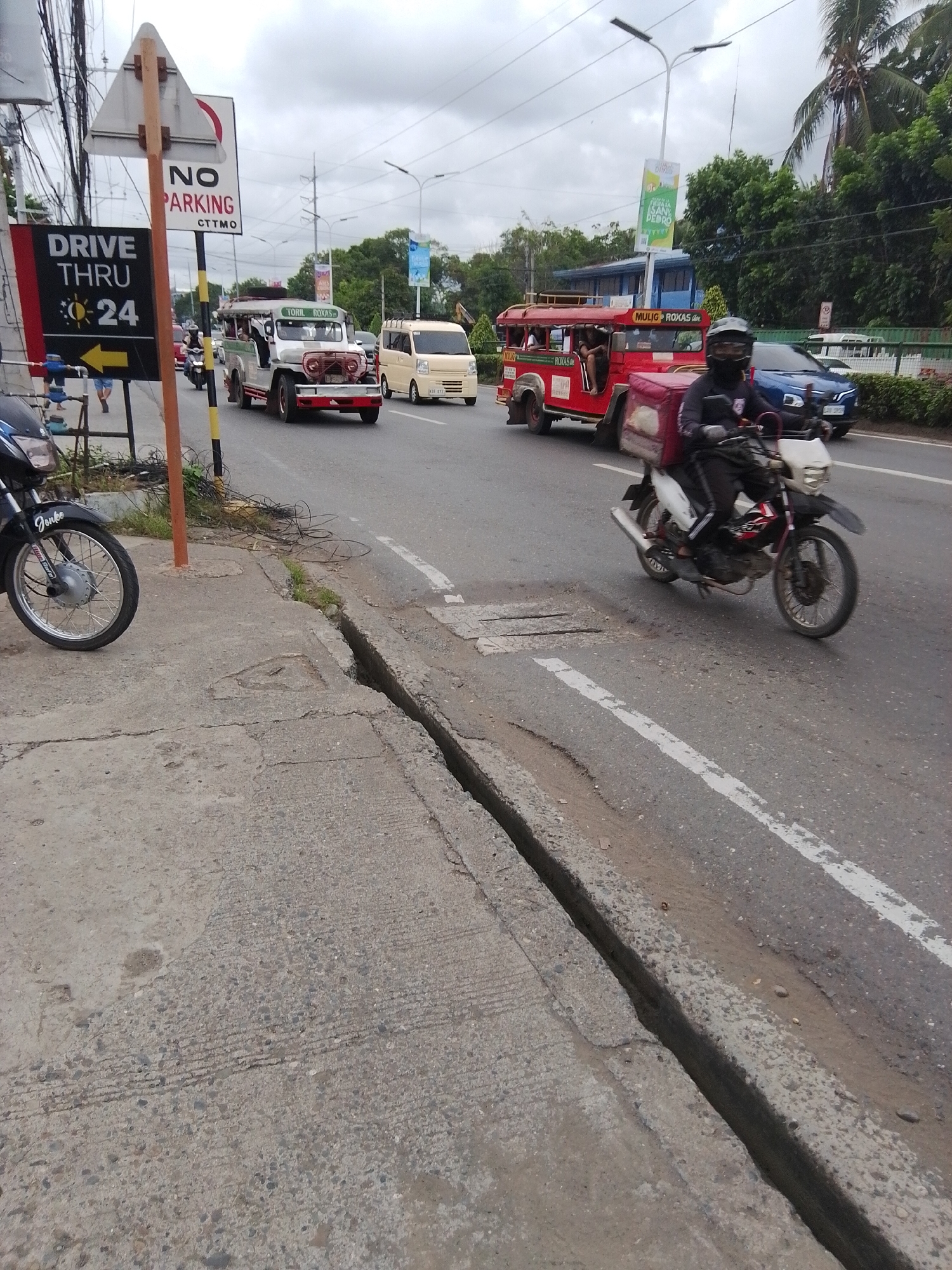 In Davao City, Philippines, a street scene unfolds with multiple vehicles and pedestrians. On the left side of the road, a motorcycle is parked on the sidewalk, facing away from the camera. Further down, a "DRIVE THRU 24" sign with a sun and an arrow pointing to the right is prominent. Next to it, a "NO PARKING" sign is posted.
The street is populated with various vehicles in motion. A white jeepney with "TORIL ROXAS" written on it is traveling in the right lane. Behind it, a beige van is also moving along. In the right lane, a red jeepney labeled "MULIO ROXAS" is visible, with passengers inside. A person on a motorcycle wearing a helmet and a dark jacket with a red backpack is in the process of overtaking the red jeepney. A blue car is also seen in the background.
The environment is a busy urban street with a cloudy sky. The road has white lane markings, and a significant crack or trench runs along the edge of the road and sidewalk on the right side of the image. Trees and buildings are visible in the distance. A few pedestrians are present near the "DRIVE THRU" sign. The overall impression is a typical day in a bustling city.