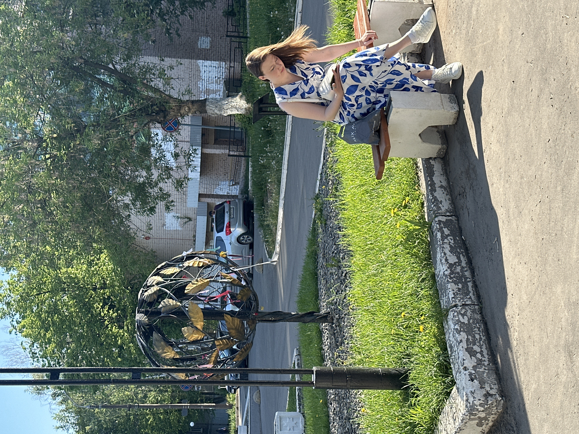 A woman sits on a bench in Izhevsk, Russia. She has long brown hair and wears a blue and white floral dress and white sneakers. She is sitting upright with her hands in her lap and her legs slightly bent. The bench is made of wood and concrete.
Behind the bench is a curb, grass, and a metal sculpture in the shape of a tree with leaves. Beyond the sculpture is a paved area, a parked silver car, and a multi-story brick building. A traffic sign is visible on the side of the building. The scene appears to be outdoors on a sunny day.