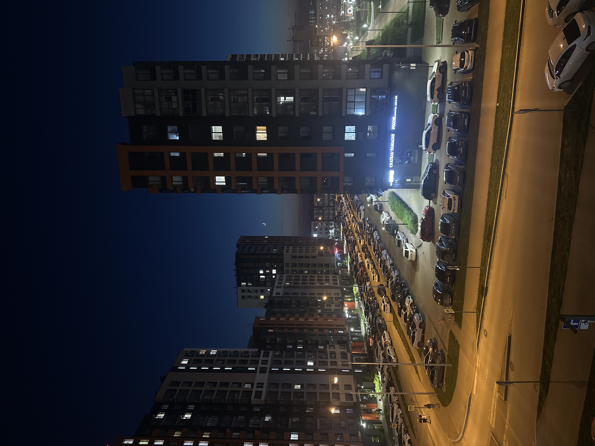 The image shows a nighttime scene in Perm, Russia, likely a residential or mixed-use area. Several tall apartment buildings are visible, illuminated by interior lights and streetlights. A long row of parked cars lines the street adjacent to the buildings. Streetlights cast a yellow glow on the road. The sky is dark, transitioning from blue to darker shades. A crescent moon is visible between the buildings. A sign that reads "Chateau Tamagne" can be seen on a building.