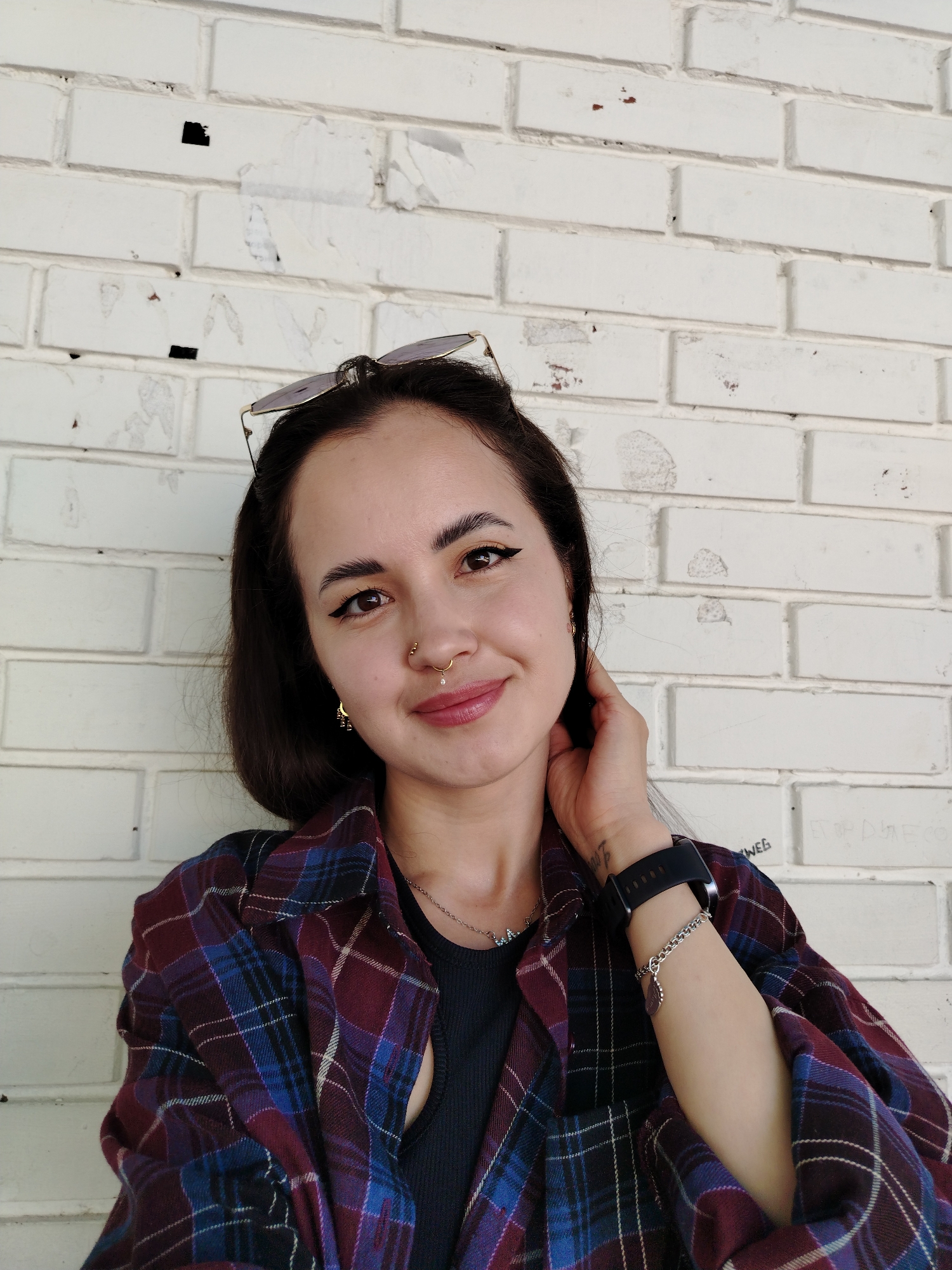 A female individual is depicted in a head-and-shoulders portrait against a white brick wall in Noviy, Russia. The individual, appearing to be in her 20s or 30s, has dark hair, styled with sunglasses positioned on her head. Her facial features include defined eyebrows, winged eyeliner, and multiple gold-toned piercings: a septum ring with a clear dangle, a ring on the left nostril, and a philtrum piercing. She wears gold-toned hoop earrings, a silver chain necklace, a black smartwatch, and a silver charm bracelet on her right wrist. Her attire consists of a black undergarment beneath an open, predominantly maroon and blue plaid shirt. She exhibits a subtle smile, looking directly into the camera, with her left hand positioned behind her head. The background brick wall shows signs of wear, including torn paper remnants, dirt, and minor discoloration.