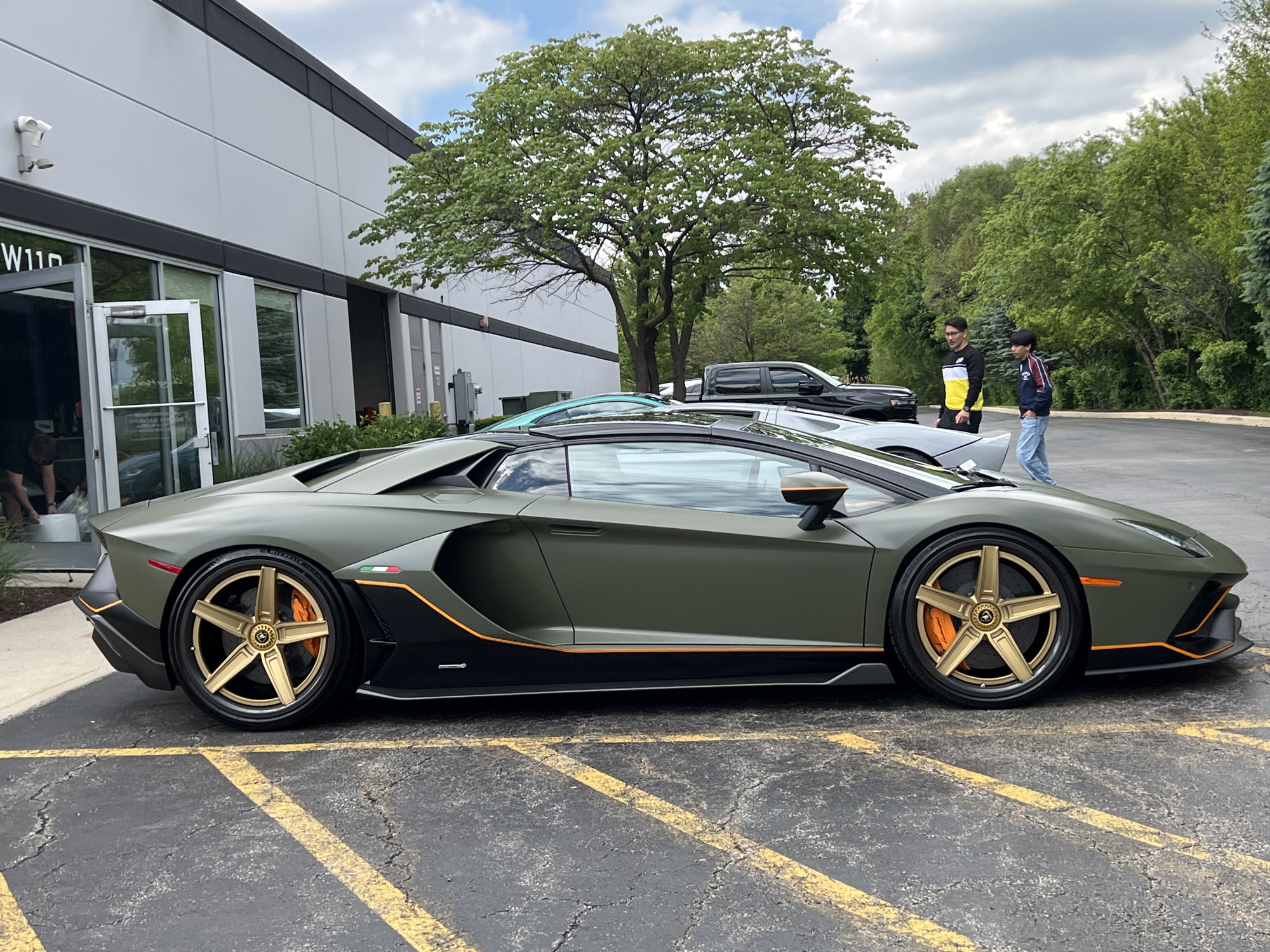 Here is a description of the image:
The image showcases a Lamborghini Aventador, painted in a matte olive green color with gold wheels and orange brake calipers, parked on a cracked asphalt surface with yellow painted parking lines. The car features black accents along the lower body and orange pinstriping.
In the background, there's a modern, gray building with glass doors and windows, with a security camera visible on the exterior. Behind the Lamborghini, another car is parked, along with a black pickup truck. Two people stand near the other car, with one wearing a black and yellow jacket. Trees and foliage fill the background, creating a natural backdrop.
The location appears to be a parking lot outside of a commercial or industrial building.