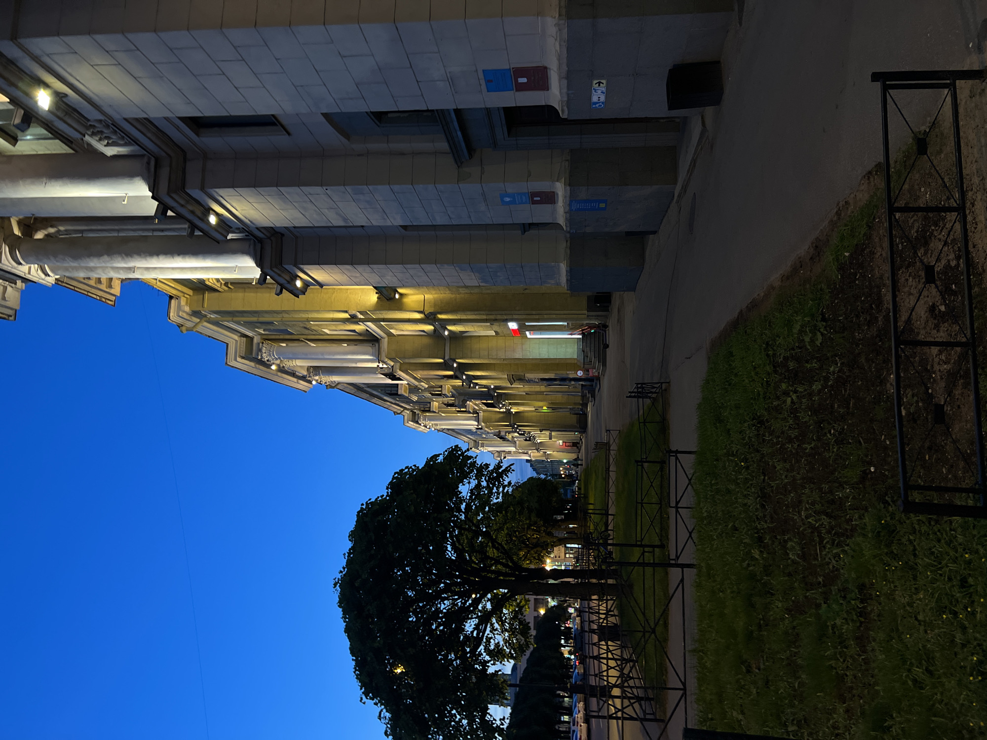The image shows a sidewalk and a building facade in the city of Unknown, Unknown at dusk. The sky is a deep blue, indicating twilight. The building is a large, imposing structure with classical architectural elements, including columns and a light-colored stone facade. Lights are illuminated on the building, casting a warm glow on the facade and the sidewalk. A row of trees lines the street in front of the building, and a black metal fence separates the sidewalk from a grassy area. There are a few cars parked on the street and a trash can is visible next to the building. There are no people or animals visible in the frame.