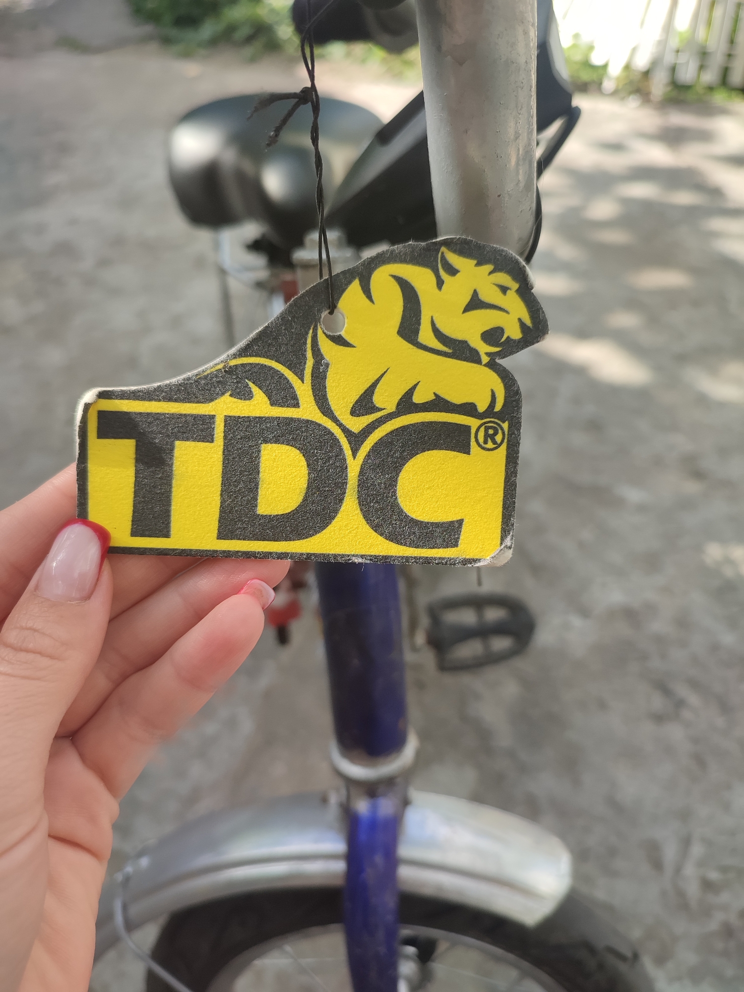 A person's hand, with manicured nails painted with a light pink polish and French tips, is holding a yellow air freshener tag. The tag is rectangular with rounded edges and has black lettering and graphics. The main graphic features a yellow tiger-like animal with black outlines, positioned above the word "TDC" in large, bold black letters. A registered trademark symbol "®" is visible to the right of the "C". The tag is attached to the handlebar of a bicycle by a black string. The bicycle is blue and silver, with a black seat and pedals visible. The background is out of focus, showing a paved surface, some greenery, and a white fence. The lighting suggests it is daytime, likely sunny, with shadows visible on the ground. The scene appears to be outdoors, possibly in a residential area in Shyshaky, Ukraine, given the provided location context. The image focuses on the air freshener and the hand holding it, implying the object is being presented or examined.