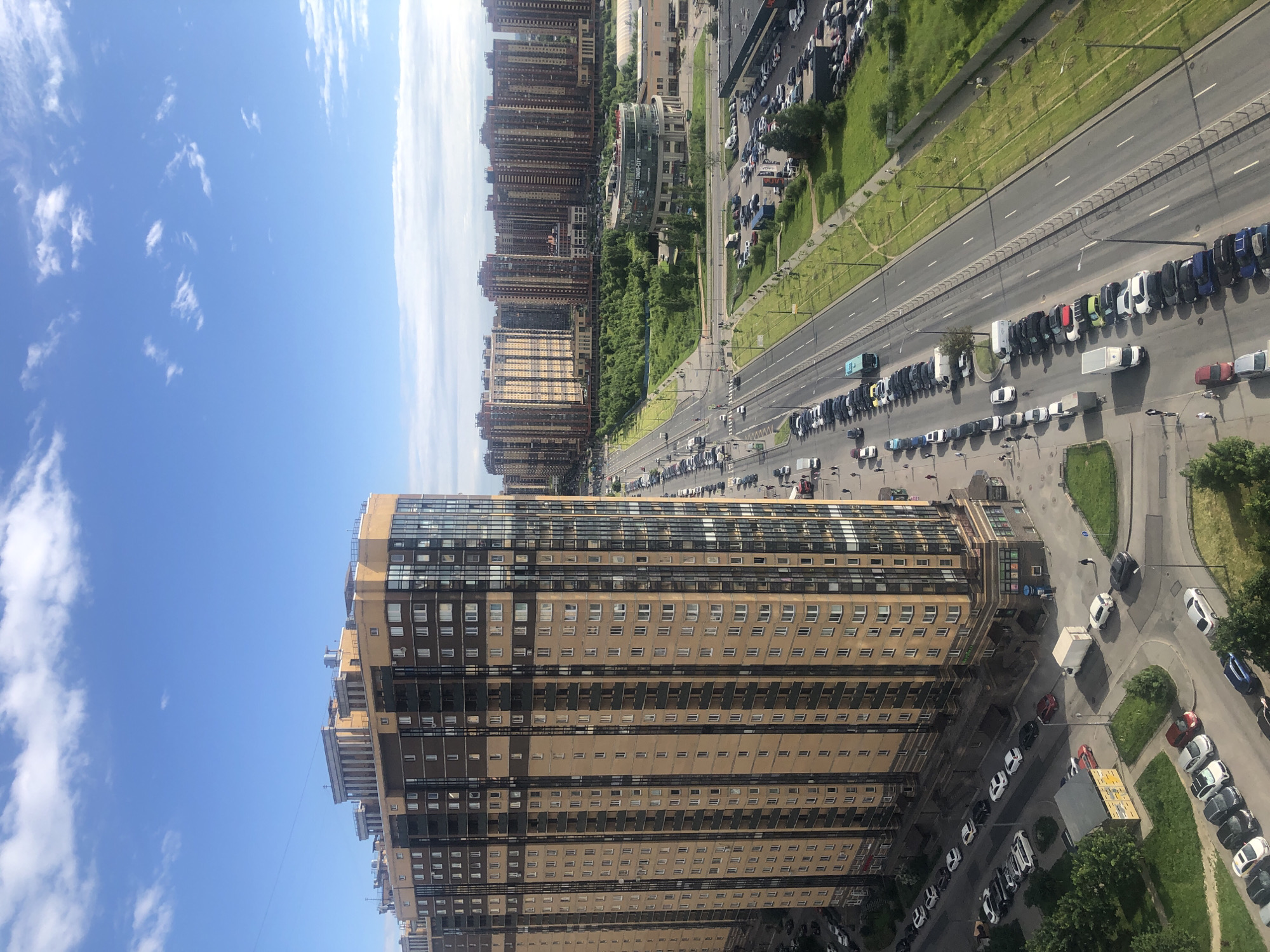 An eye-level, high-angle, long shot captures a dense urban landscape under a partly cloudy blue sky. Dominating the left side of the frame is a tall, modern apartment building, its facade a combination of beige panels and dark-framed windows, punctuated by balconies. To the right, a multi-lane road stretches into the distance, lined with parked cars and a grassy median. Beyond the road, a cluster of similar apartment buildings forms a backdrop. In the middle ground, a circular building with glass walls stands amidst lush green trees, labeled "TOKYO-CITY". A few people are visible walking on sidewalks and near the road. The scene appears to be a typical suburban or outer-city residential area with significant infrastructure.