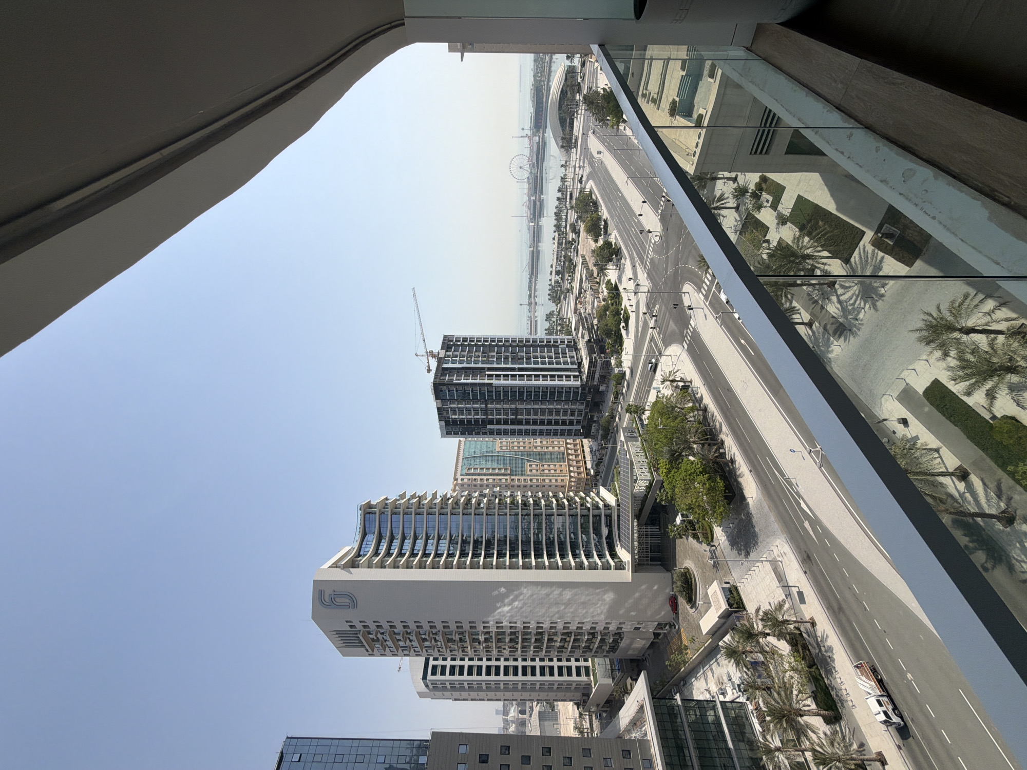 The image captures a high-angle view of a modern urban scene in Lusail, Qatar, under a clear blue sky. In the foreground, a balcony railing made of glass and metal overlooks a wide, empty street intersection with palm trees lining the pavements. Several multi-story buildings, characterized by their contemporary architecture with glass facades and sleek designs, dominate the mid-ground. One prominent building features a distinctive logo with the numeral "6". Further in the distance, a waterfront area is visible, featuring a large Ferris wheel, a curved structure, and other buildings. A construction crane is also present, indicating ongoing development. A white truck is seen on the street, and there is a red car parked near the entrance of one of the buildings. The overall environment suggests a well-planned, modern city district with a focus on open spaces and high-rise architecture.