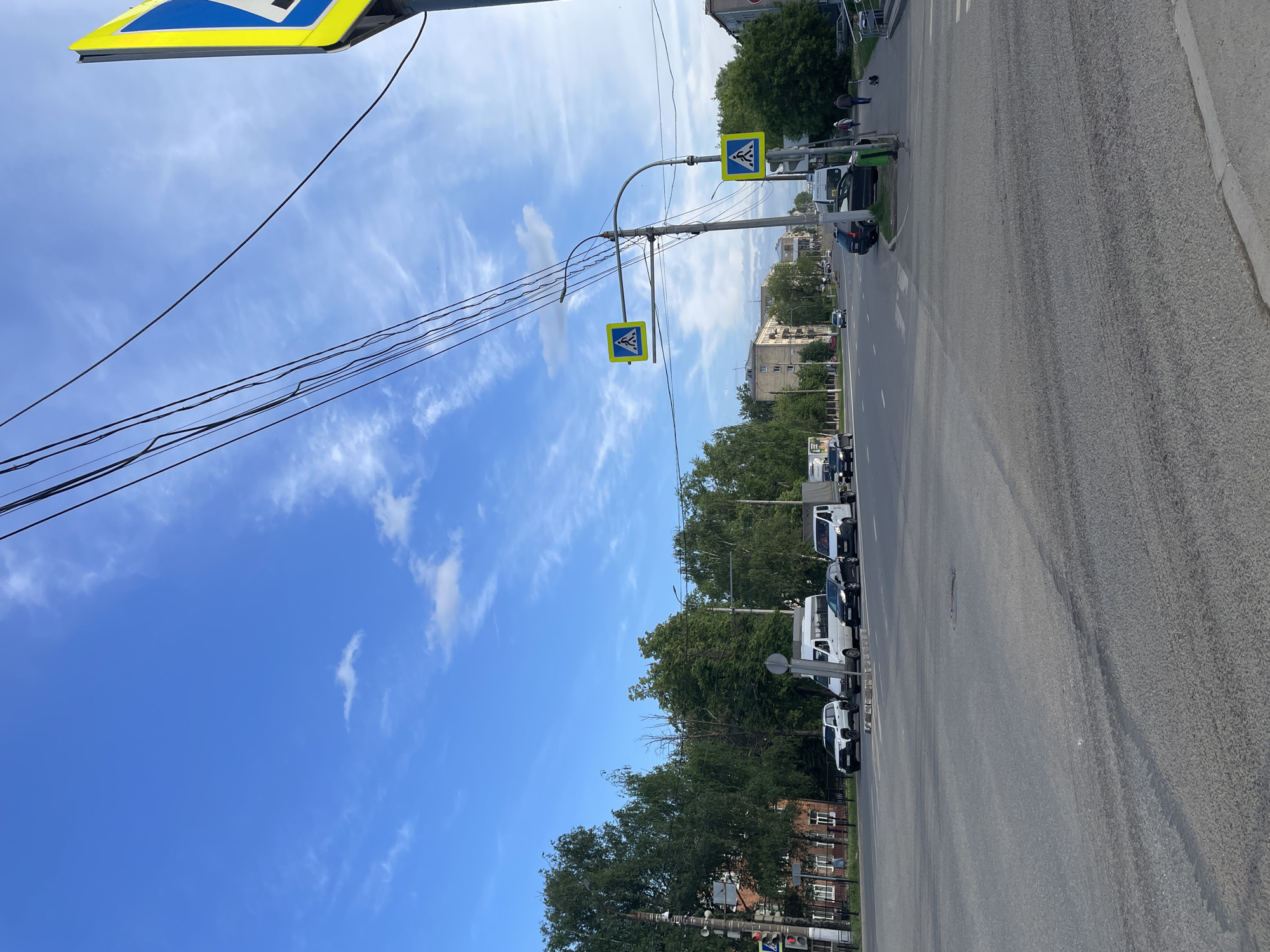 The image depicts a street scene in Izhevsk, Russia, characterized by a clear blue sky with scattered clouds. A prominent feature is a crosswalk signpost supporting two signs with a pedestrian crossing symbol. Several black electrical wires extend from the top left corner of the image towards the signpost. On the right side of the frame, there is a multi-lane road with a mix of parked and moving vehicles, including cars and vans. Trees line the street, and buildings are visible in the background, indicating an urban environment.
