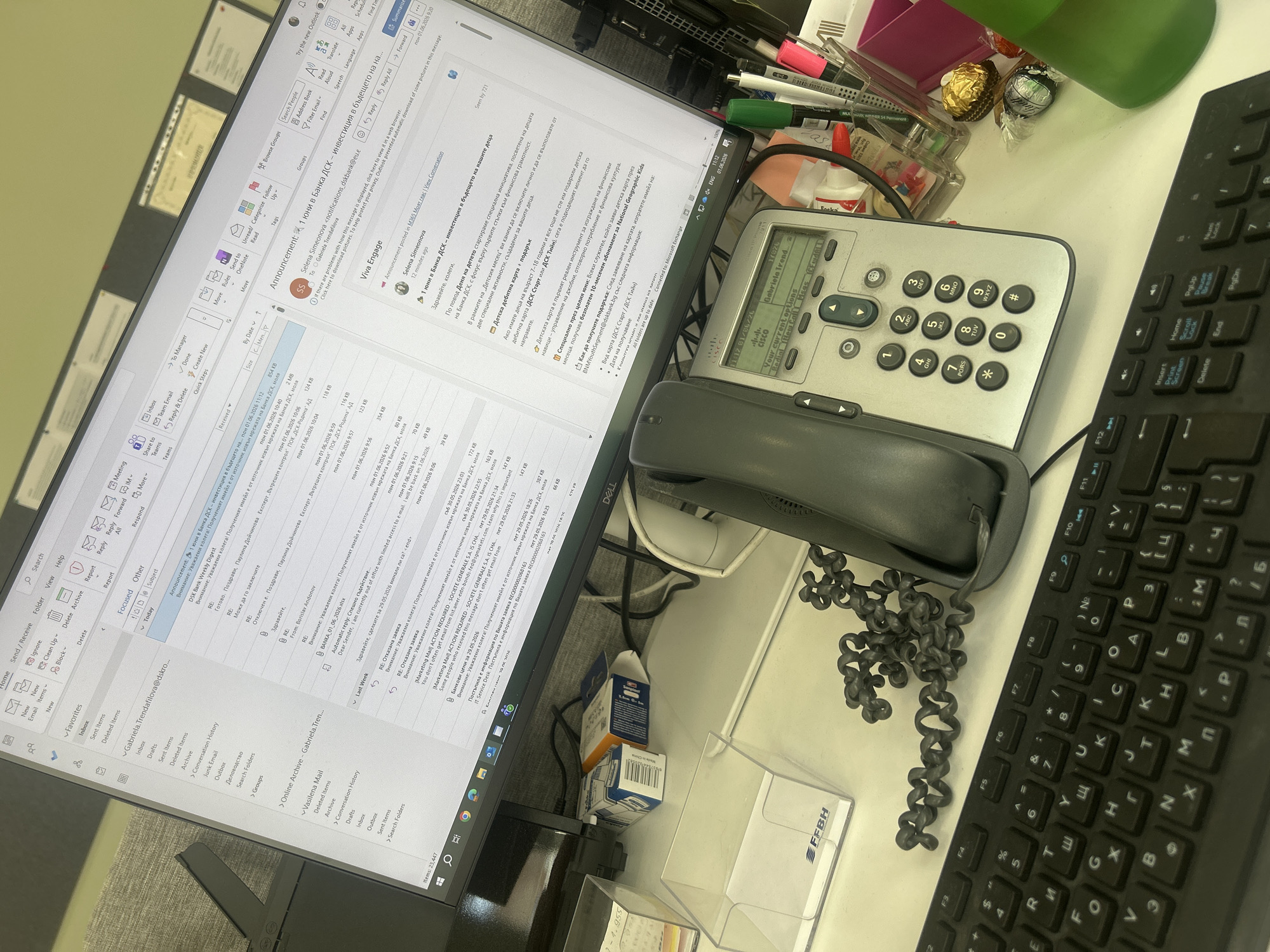 Here is a detailed description of the image:
**Scene:** A close-up, high-angle shot of a cluttered office desk.
**Objects:**
* **Electronics:**
* A Dell monitor displaying the Microsoft Outlook email application. The inbox is visible, with numerous emails.
* A Cisco desk phone with a handset resting on the base. The phone's display shows current calls/interceptions.
* A black keyboard with Cyrillic script.
* **Office Supplies:**
* Pens and markers.
* A clear acrylic business card holder.
* **Food and Drink:**
* Chocolates (Ferrero Rocher and Lollipops).
* A green bottle.
* **Other:**
* A small white box.
* Telephone cord.
**Computer Screen:**
* **Email:** The Outlook application shows the inbox. Numerous emails are visible, with subjects in Bulgarian, indicating business communications within a Bulgarian bank.
* One email mentions "1 юни в Банка ДСК - инвестиция в бъдещето на нашите деца" (June 1st in DSK Bank - investing in the future of our children).
**Inferences and Location:**
* **Location:** The use of Cyrillic script on the keyboard and the subject line suggests Sofia, Bulgaria. The frequent mention of "Банка ДСК" (DSK Bank) in the emails further strengthens this location, as DSK Bank is a prominent bank in Bulgaria.
* **Occupation:** The desk setup suggests the person works in an office environment, likely at DSK Bank, dealing with electronic communications and telephone calls.