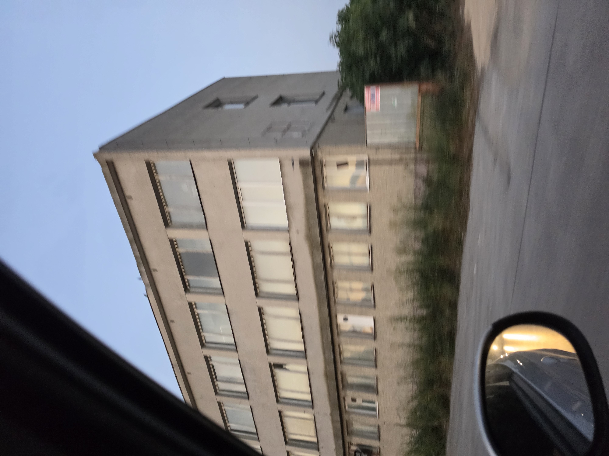 An exterior, medium-long shot captures a three-story, Brutalist-style building situated in Topoli, Bulgaria. The building is constructed of light-colored brick and features a series of uniformly spaced rectangular windows on each level, with some windows appearing reflective. A dark, curved object, likely part of a vehicle's interior, occupies the left side of the frame, obscuring a portion of the sky and the upper left corner of the building. The foreground shows a paved road with blurred vegetation and a side-view mirror reflecting lights. The sky is a clear, pale blue, indicating it is either early morning or late evening. The overall scene suggests the building is possibly an office or industrial complex.