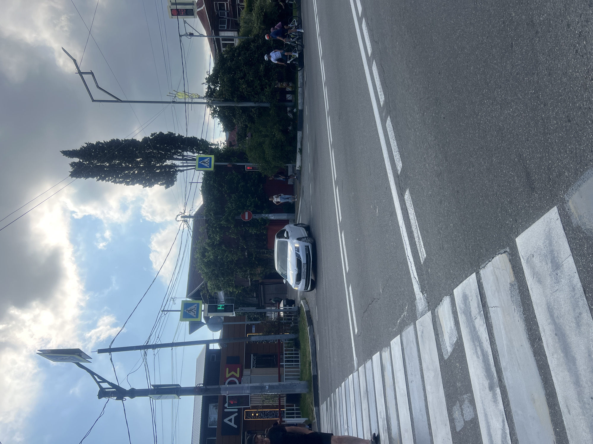 The image shows a street intersection in a city, likely during the daytime. A white car is stopped at a pedestrian crossing, waiting for traffic. Several people are visible in the scene, including pedestrians waiting to cross the street and cyclists riding past. Traffic signals are visible, with one showing a green light. The environment includes buildings, trees, and streetlights, indicating an urban setting. The sky is partly cloudy.