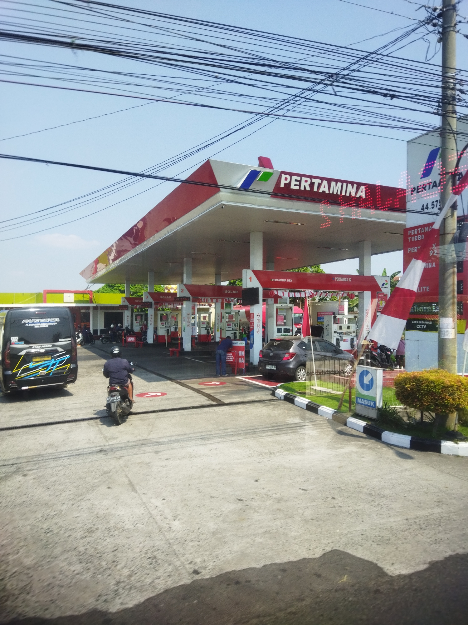 This image depicts a Pertamina gas station located in Begajah, Indonesia. The gas station features a red and white canopy supported by white pillars. Several fuel pumps are visible, with one labeled "PERTAMAX 92". A dark gray car is parked at a pump, and a person in a blue shirt is standing near it. A motorcycle is also present, with a rider on it heading away from the camera. To the left, a black van with yellow and blue graphics is parked. The ground is paved concrete with painted directional arrows. In the background, there are buildings and trees, and the sky is clear blue. Overhead, a dense network of black electrical wires crisscrosses the sky. The overall scene captures a typical day at a busy gas station.