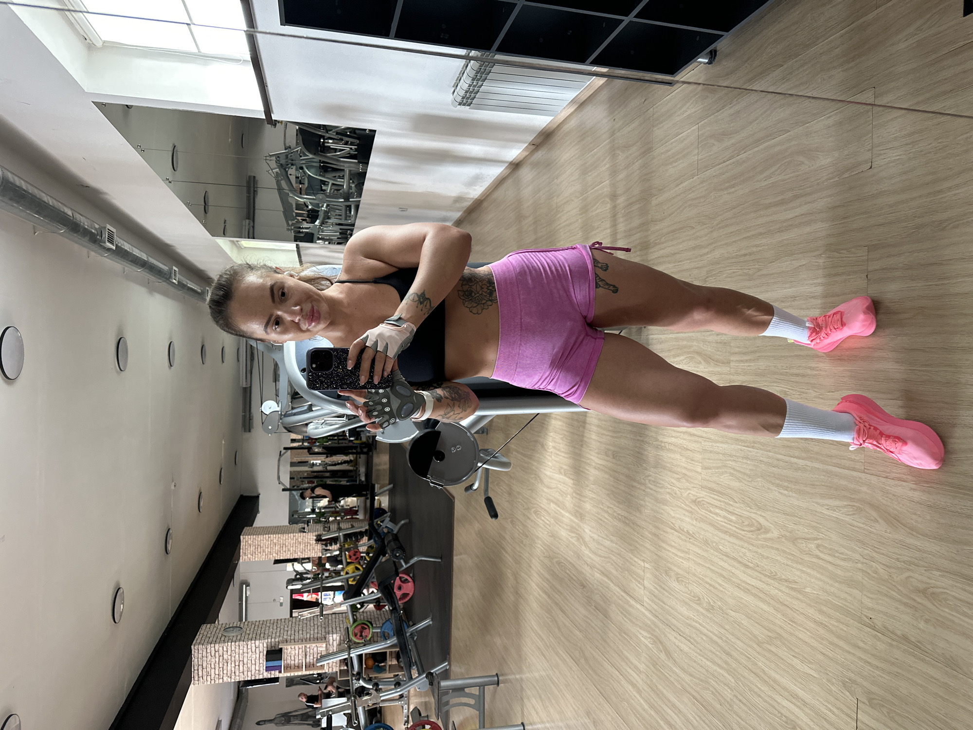 A woman is taking a selfie in a gym. She is wearing a black sports bra, pink shorts, white socks, and pink athletic shoes. She has on grey workout gloves and is holding a smartphone in her right hand. There are tattoos visible on her left arm and thigh. The gym has wooden floors and is equipped with various weight machines and benches. Mirrors line one wall, reflecting the gym equipment and a radiator. The woman's reflection is prominent in the foreground. The image appears to have been taken indoors in a gym setting, which could be located anywhere, including Yuzhnouralsk, Russia.