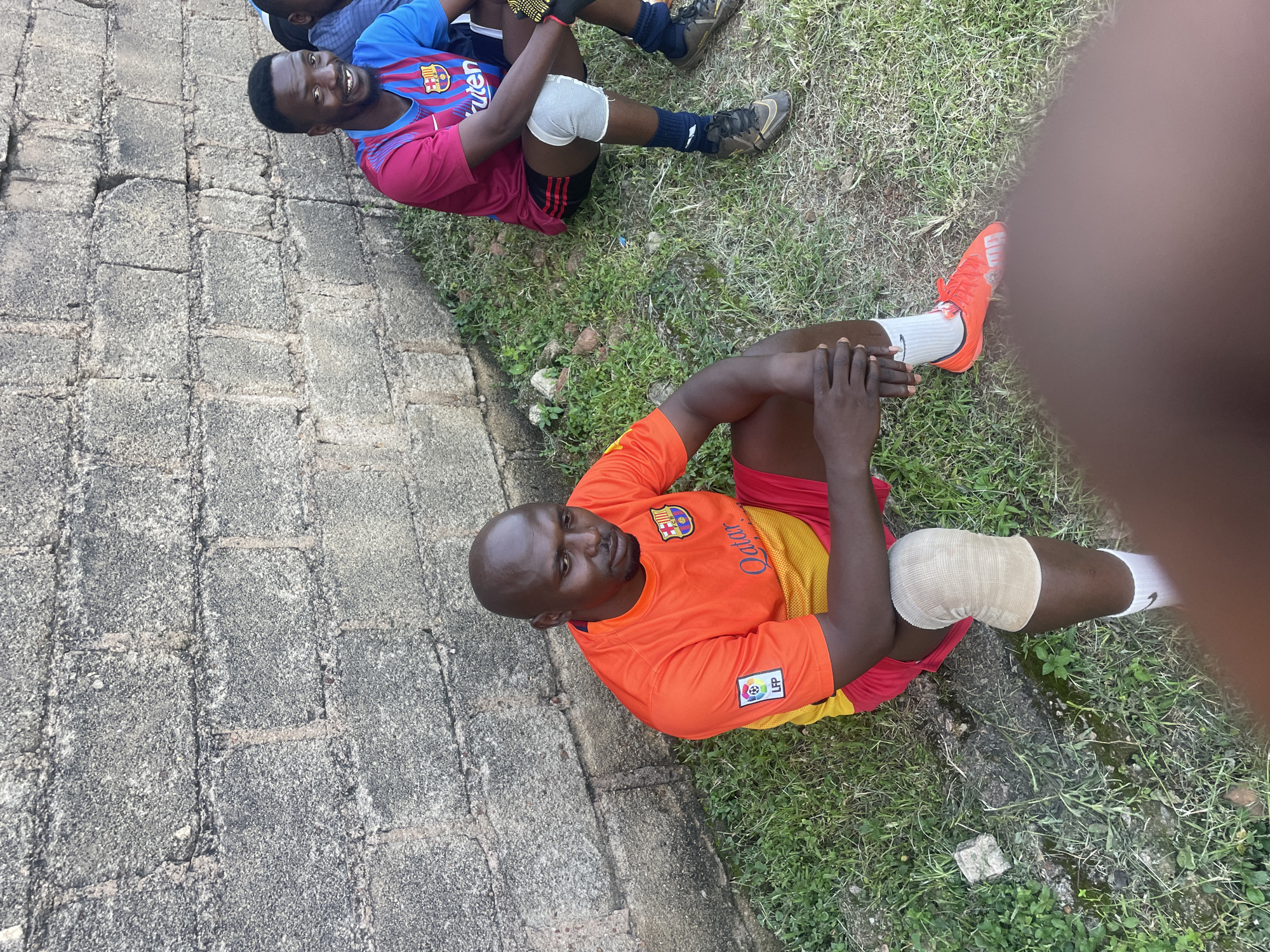 Two men are sitting on the ground next to a grey brick wall in a grassy area, possibly in Jos, Nigeria. The man in the foreground is wearing an orange and yellow soccer jersey with the "Qatar" logo and a red soccer shorts, and has a knee brace on his left leg. The man in the background is wearing a blue and red soccer jersey with the FC Barcelona logo. Both men appear to be resting, perhaps during or after a sports activity. The environment suggests an outdoor urban setting with a weathered wall and a patch of green grass.