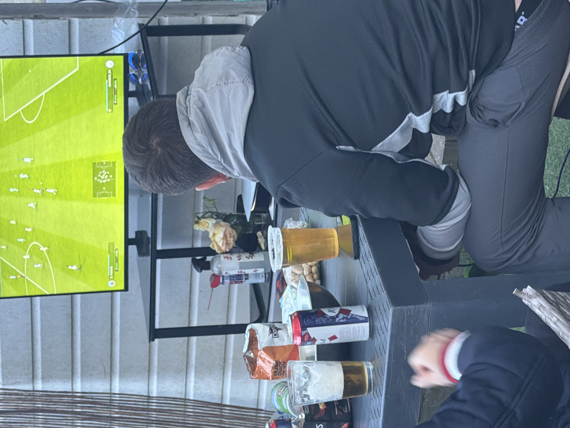 The scene depicts an outdoor setting, likely a garden or patio in Maidstone, United Kingdom. An adult male, with short dark hair, is seated with his back to the camera, wearing a dark grey jacket with light grey accents and a hood, along with dark grey trousers. He is positioned in front of a flat-screen television displaying a football video game with a bright green pitch, white-clad players, and an overhead perspective.
To the left of the male, a partial view of another individual's arm and upper torso is visible, wearing a dark blue jacket with red and white striped cuffs. Both individuals are seated at a dark grey, rectangular outdoor table.
On the table, from left to right, are: a half-empty pint glass containing beer, a partially visible dark-labeled bottle, an upright red, white, and blue can (partially obscuring "104" branding), an open bag of "Doritos Golden Crunch" crisps, and a silver bowl filled with peanuts or similar nuts. Further to the right, a clear plastic cup also holds a golden liquid, likely beer, and a black smartphone lies face-down on the table. Behind the snacks, a clear spray bottle with a white nozzle and a red applicator straw is present, next to a small container holding dried, cream-colored flowers.
The television is placed on a black stand/table. Behind the TV stand, a blue object, possibly a gaming controller, is visible. The background features light-colored horizontal siding, suggestive of a building exterior, and vertical bamboo screening on the far left. Green foliage is partially visible at the bottom of the frame.