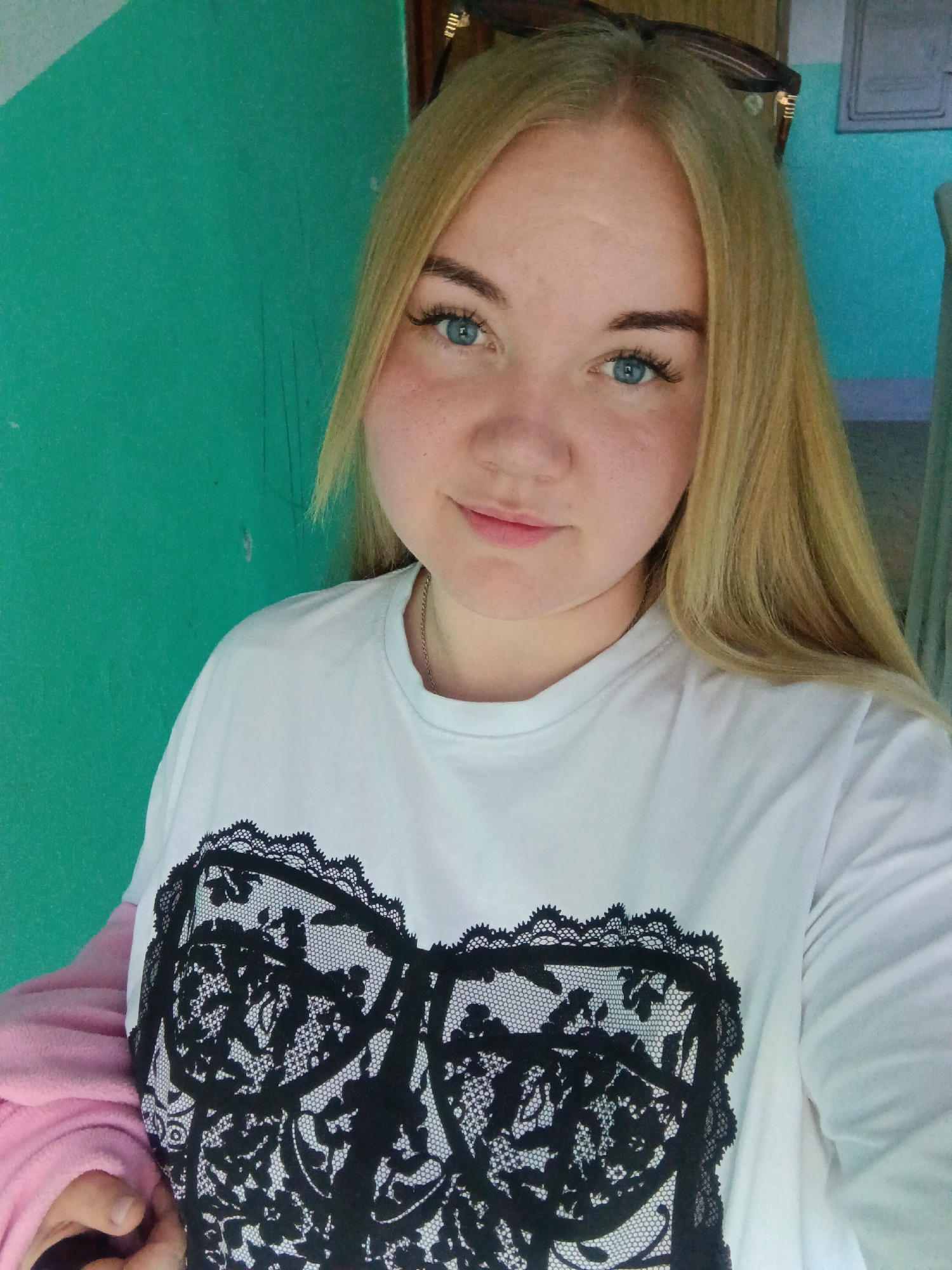 A young woman is depicted in a close-up, indoor selfie, set against a vibrant teal or turquoise wall. She has long, blonde hair, blue eyes, and fair skin with visible freckles across her nose and cheeks. Her eyebrows are well-defined, and she has long eyelashes. She wears dark sunglasses pushed up onto her head.
The woman is wearing a white, long-sleeved t-shirt featuring a prominent black graphic on the chest designed to resemble a lace bra or corset. A thin, silver-colored chain necklace is visible around her neck. She holds a soft, pink garment, possibly a jacket or blanket, in her left arm/hand at the bottom left of the frame. She looks directly at the camera with a soft, pleasant expression and a slight smile.
The background shows hints of other indoor structures or walls, slightly out of focus, suggesting she is inside a building, perhaps a home or public space in Chaikovsky, Russia. The lighting appears to be natural, indicating it is likely daytime. There is no visible text in the image.