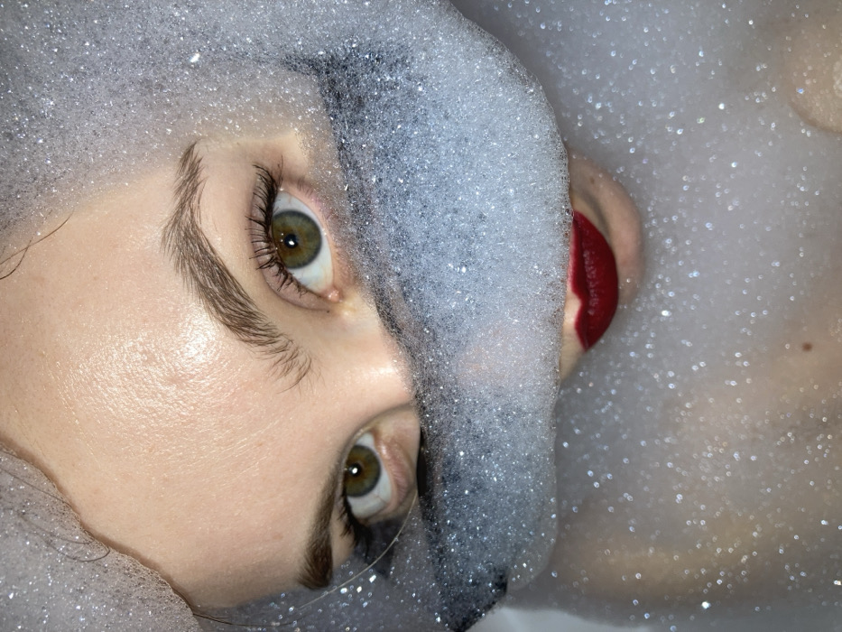A woman lies surrounded by white suds. The camera focuses close on her face, tilted at an angle. Her eyes, a hazel-green, stare upwards. Her skin is pale, reflecting light with a sheen. A dark, undefined shape cuts vertically across her face from her hairline. Deep red lipstick contrasts sharply against the pale skin and white bubbles. More suds fill the background, creating a sparkling, almost ethereal effect. A hint of bare skin and a small mole are visible on the right.