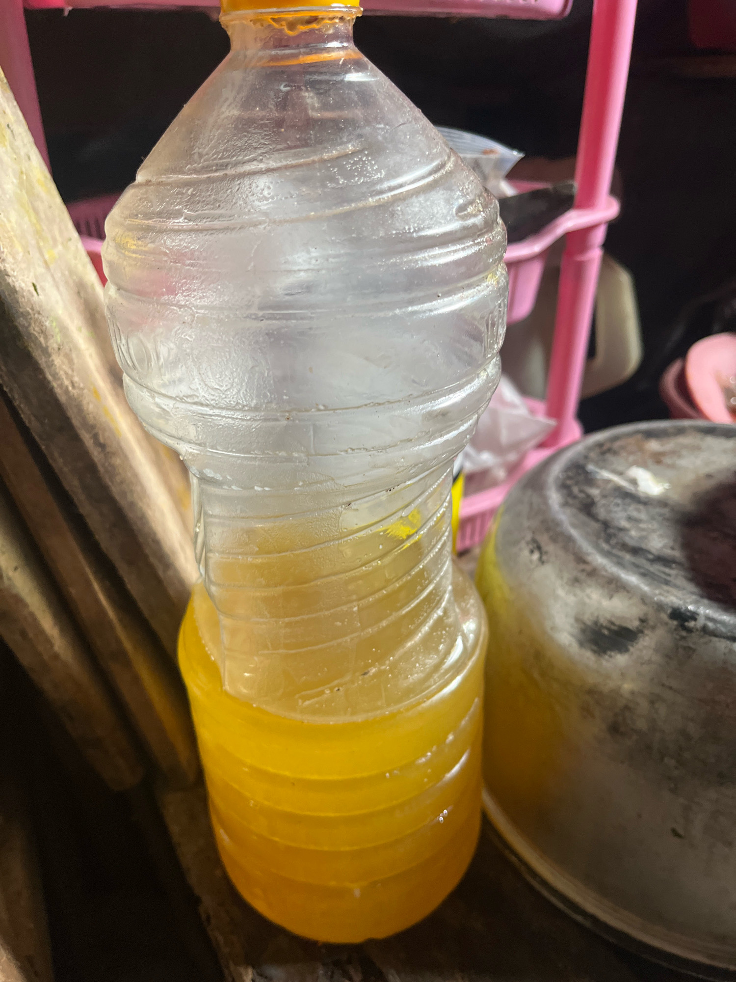 In Sidomulyo, Indonesia, a close-up, low-angle shot reveals a plastic bottle filled with a yellow liquid and ice. The bottle is positioned in the center of the frame, with its top obscured. The yellow liquid occupies the bottom half of the bottle, while the top half is filled with ice. To the left of the bottle, a wooden object with a rough texture is visible. In the background, a pink plastic shelf unit stands out, with some out-of-focus items on its shelves. To the right of the bottle, a large, metallic container with a worn, mottled surface is present. The lighting appears to be artificial and casts shadows, creating a somewhat moody atmosphere.