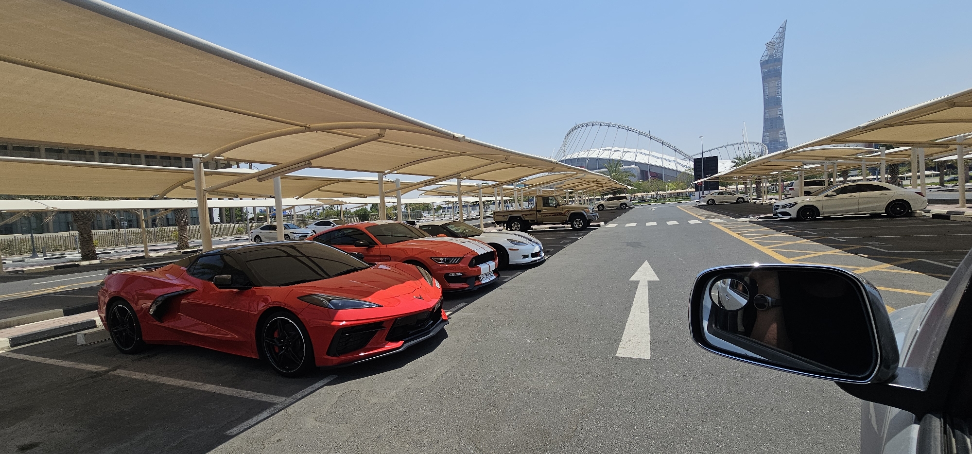 The image shows a parking lot with a canopy of beige fabric shades supported by white pillars. Several cars are parked under the shades and in the open lot. In the foreground, a red sports car is parked on the left. To its right, an orange sports car with white racing stripes is parked, followed by a white sports car. Further down the lot, a beige pickup truck is visible, along with other cars, including a white sedan. In the background, a large stadium structure with a distinctive arched roof is visible, as well as a tall, slender skyscraper. The scene is set under a clear blue sky. This location is likely the Khalifa International Stadium precinct in Al Rayyan, Qatar, given the architecture of the stadium and the prominent skyscraper which is the Aspire Tower. The image is taken from a vehicle, as indicated by the driver's side mirror in the lower right corner.