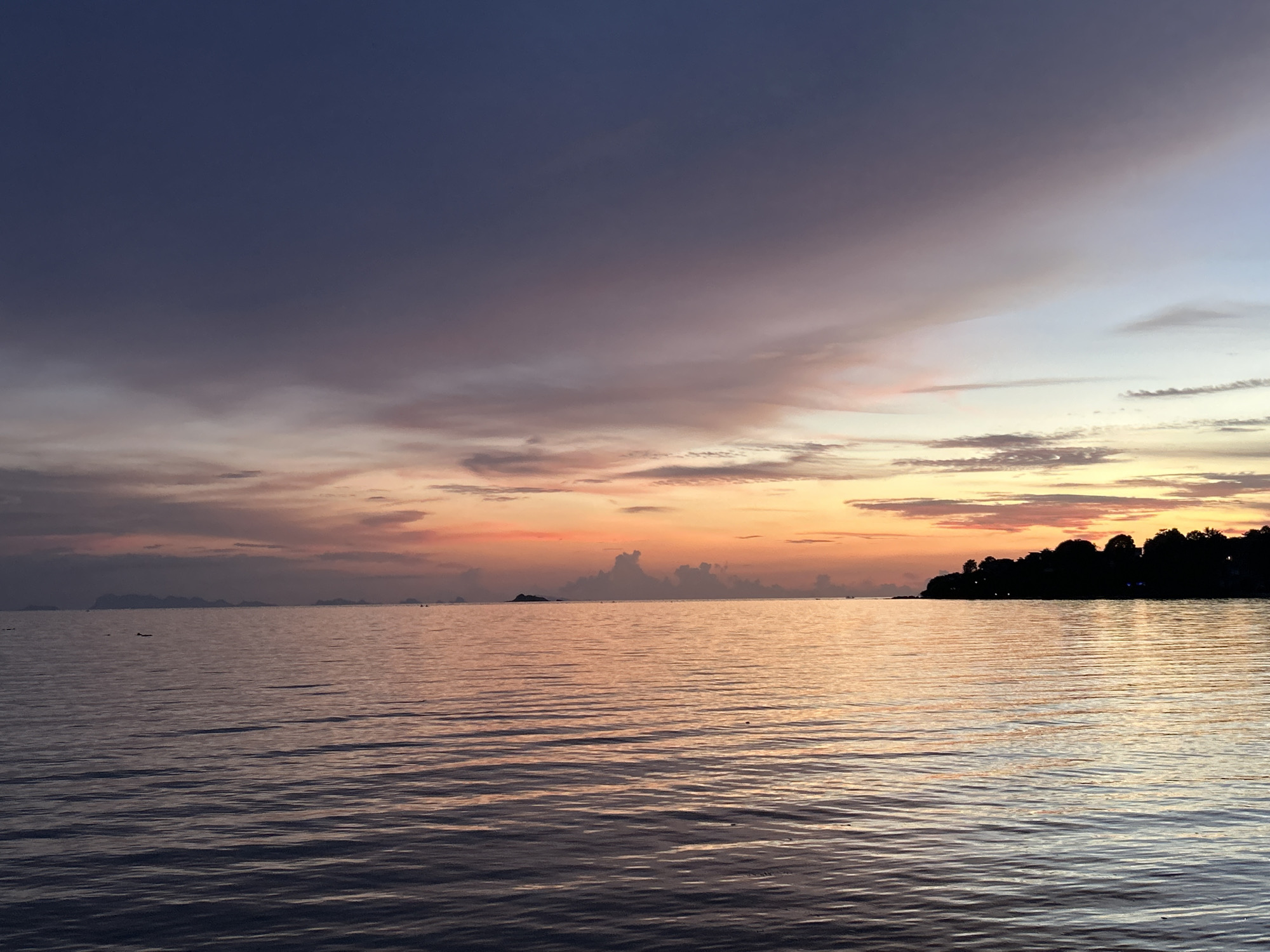 This is a serene sunset image taken from the shore of Ko Pha-ngan District, Thailand. The sky is a gradient of colors, transitioning from deep blue and purple at the top to vibrant orange and pink near the horizon, indicative of twilight. Wispy clouds are scattered across the sky, catching the fading sunlight. The sea is calm, with gentle ripples reflecting the colors of the sky and a hint of light on the water's surface. In the distance, the silhouette of a tree-lined coastline with a few scattered lights suggests the presence of a settlement. The overall scene conveys a sense of peace and natural beauty. There are no people or specific objects prominently featured, focusing the composition on the landscape.