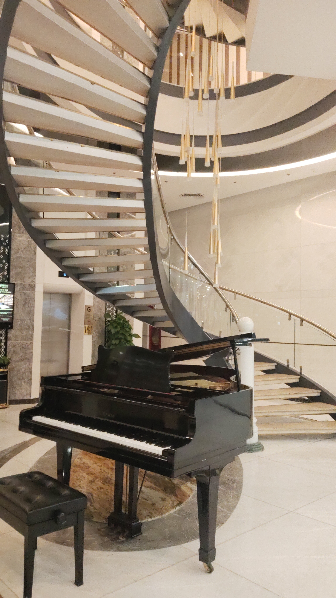 The image captures a scene in Bac Nha Trang Ward, Vietnam, featuring a grand piano alongside a spiral staircase. The piano, a black Kawai grand piano, is positioned prominently in the foreground. Next to the piano is a matching stool. The spiral staircase rises behind the piano. Above the piano and staircase, there is a decorative chandelier. The flooring is a light-colored tile. In the background, an elevator and a plant are visible. The overall setting suggests a luxurious space within a building.