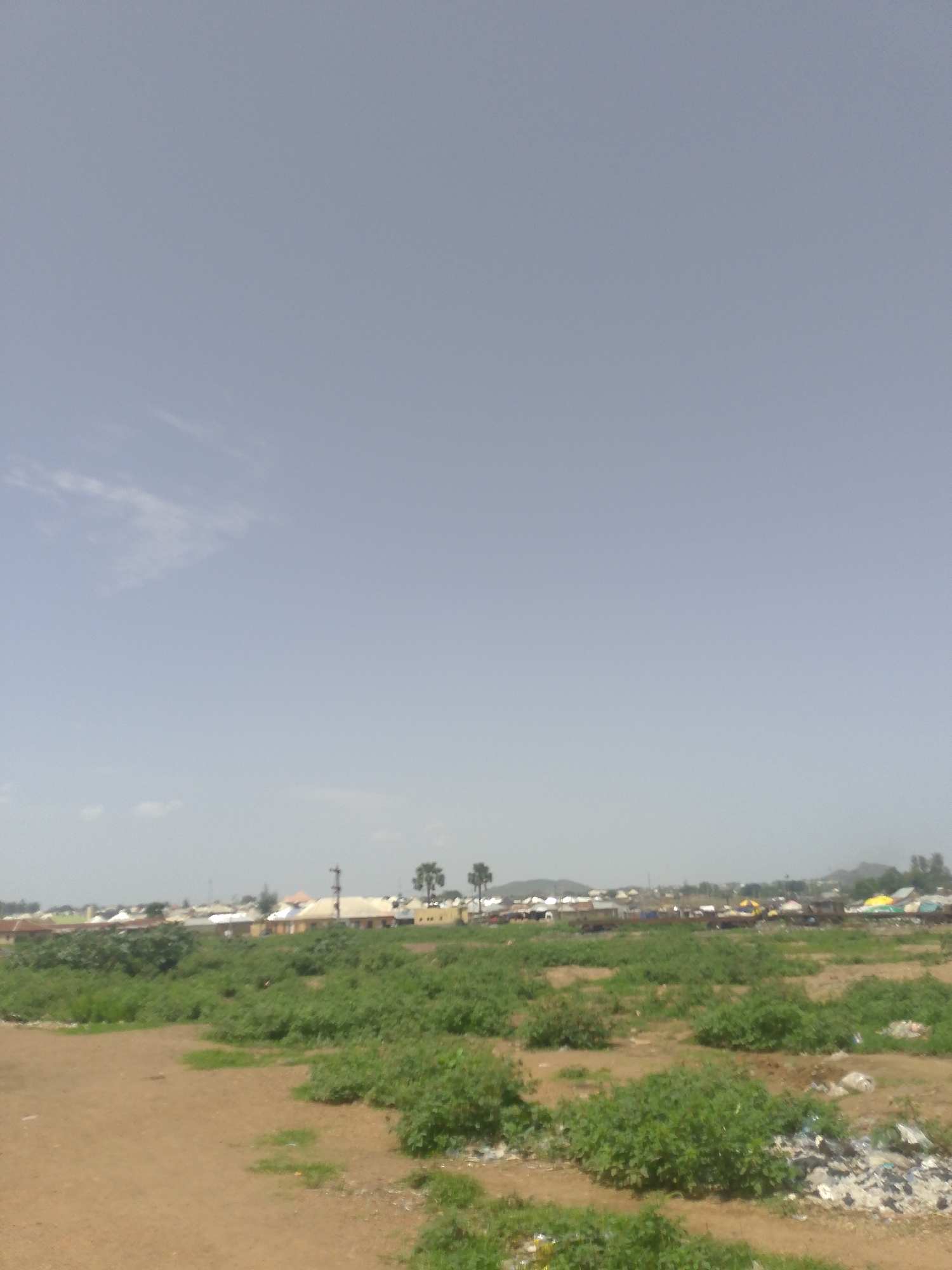 The image shows a landscape in an unidentified city. In the foreground, there is a dirt ground with patches of green bushes. Further back, a cluster of buildings and structures can be seen, with occasional palm trees interspersed. The sky is pale blue with minimal clouds. There is a small hill in the distance. At the bottom right, there is a pile of trash.