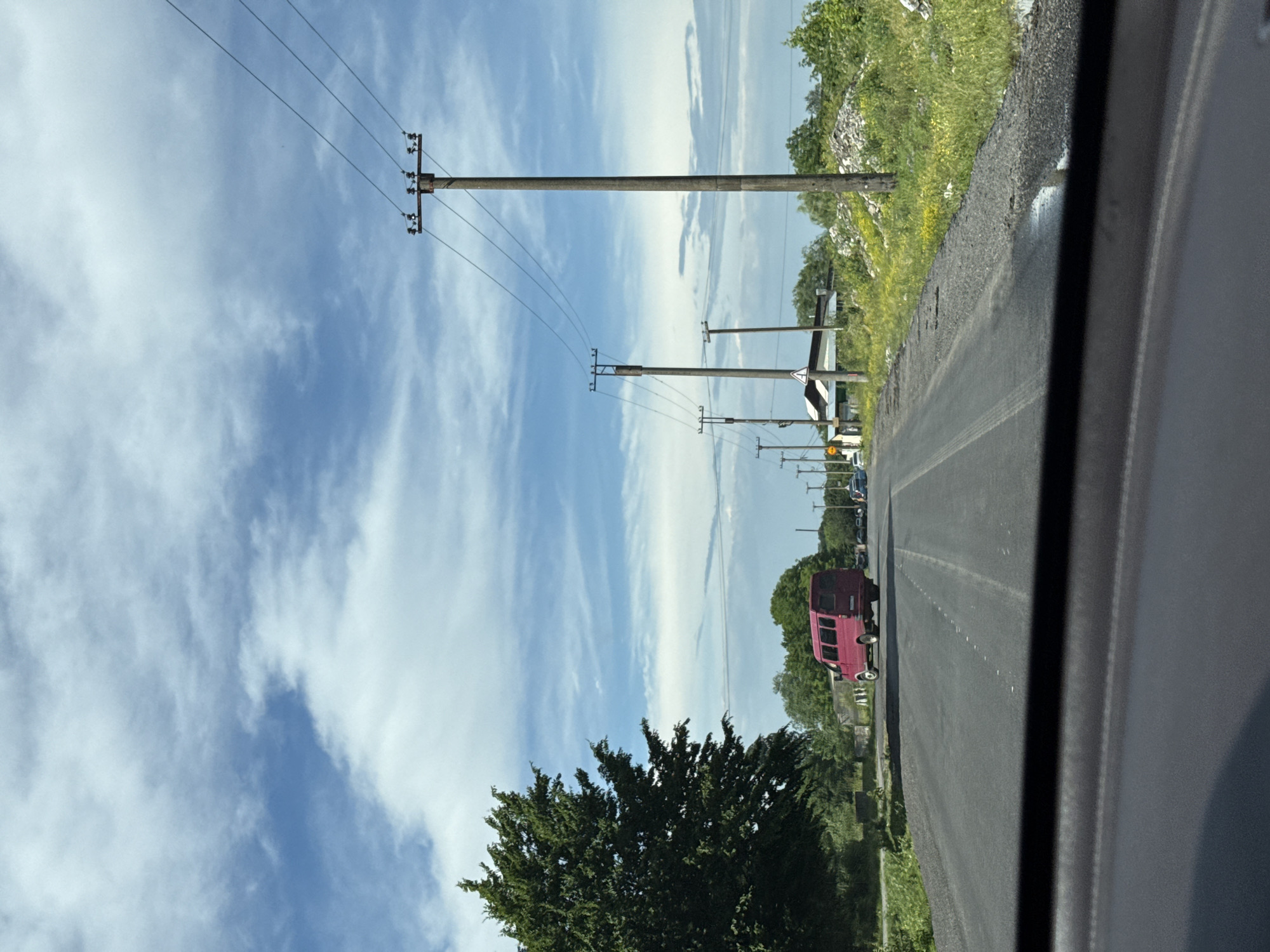 The image captures a view from inside a vehicle traveling on a paved road through a rural or semi-rural area. The sky is bright blue with scattered white clouds. On the left side of the road, a large, dense green tree stands prominently. In the distance, a red minivan is driving away from the camera. The road is lined with utility poles carrying electrical wires. On the right side of the road, there is vegetation, piles of debris or gravel, and more utility poles. In the background, houses and trees suggest a settlement. The overall impression is of a sunny day in a somewhat undeveloped area. The location is identified as the city of Alagir, Russia.