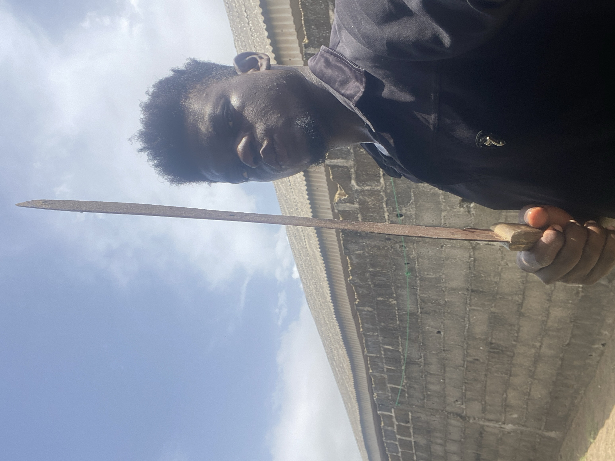An individual is holding a long metal blade, possibly a machete, with a wooden handle. The person has dark skin and short, curly hair and is wearing a dark-colored shirt. Behind the person, there is a grey brick or concrete wall. Above, there is a view of a partly cloudy sky. The location is Unknown, Unknown.