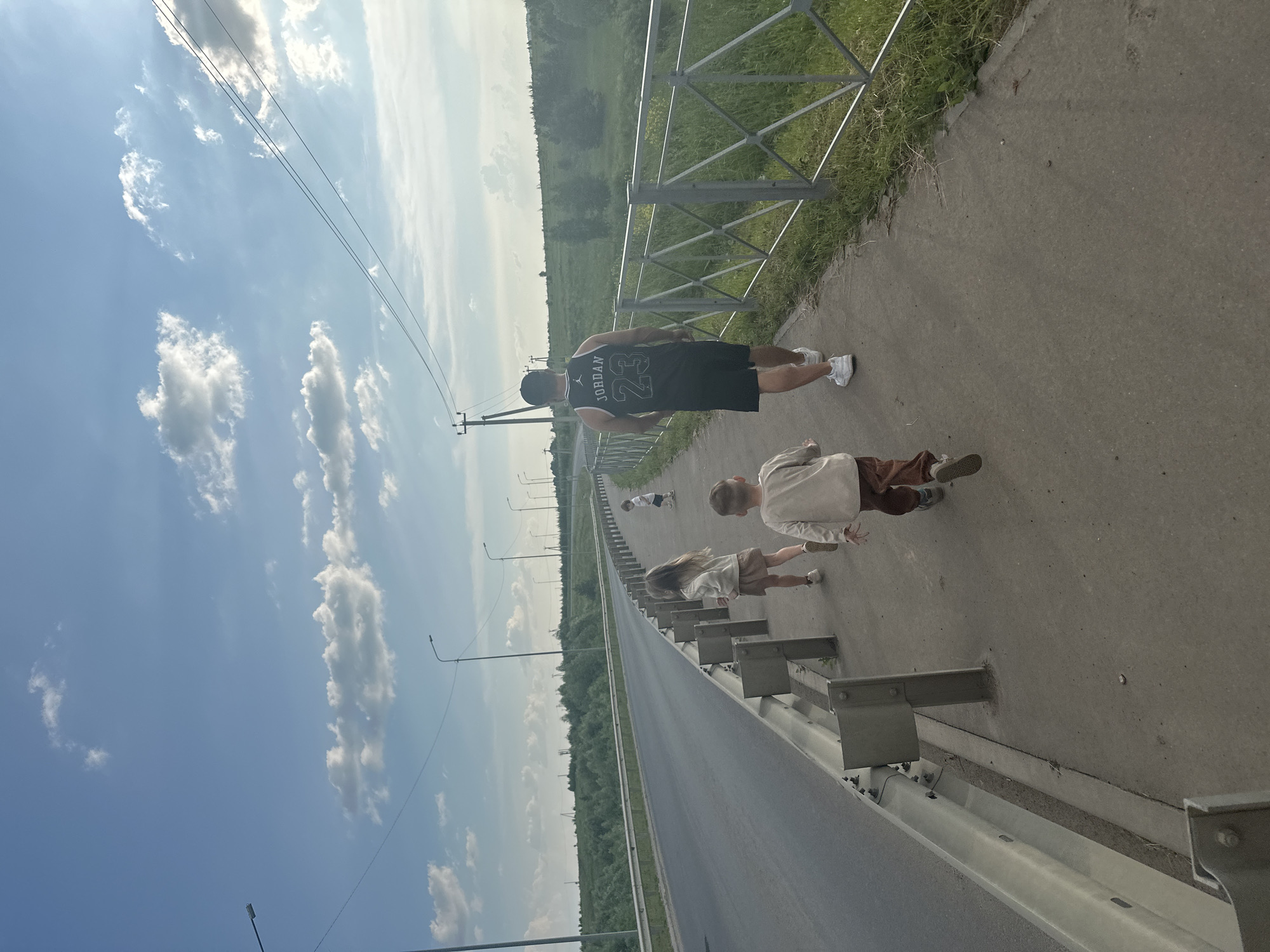 A group of four people, an adult male and three children, are walking along a paved sidewalk next to a road. The adult male, wearing a black tank top with "JORDAN 23" on the back and black shorts, is walking away from the camera. He is wearing white sneakers. To his left and slightly ahead are three children. The closest child is a boy wearing a light-colored long-sleeved shirt and brown pants, also walking away from the camera. To his left is a girl with long blonde hair, wearing a light-colored jacket and a skirt, walking in the same direction. Further ahead and to the right is another young child, partially obscured, wearing a white t-shirt and shorts. They are walking along a sidewalk bordered by a metal guardrail and a grassy embankment with trees and foliage beyond. The road to the left is empty. Overhead, the sky is blue with scattered white clouds, and power lines are visible. Streetlights are positioned along the side of the road. The scene appears to be in a semi-rural or suburban area.