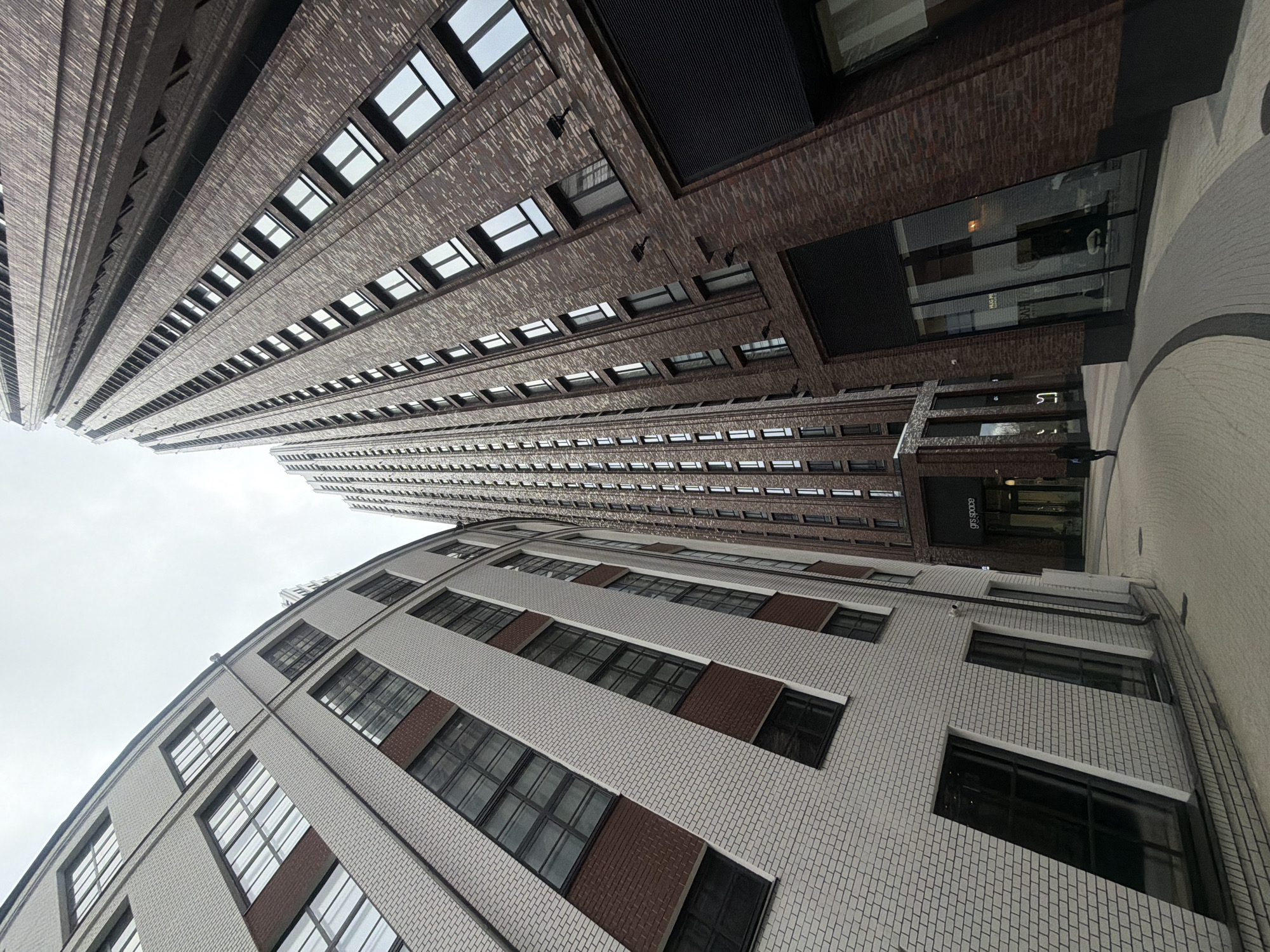 The image captures a low-angle view of buildings in Moscow, Russia, under an overcast sky. Two primary buildings dominate the scene. A taller building, constructed with dark brown bricks and featuring rows of windows, towers over a smaller, light-colored building with white brick and brown accents. The composition emphasizes the contrasting architectural styles and materials of the buildings. A sign for "gis.space" is visible on one of the buildings. The perspective is tilted, creating a dynamic and somewhat disorienting effect. The pavement is visible in the foreground, indicating the location is a street. No people are clearly discernible in the image.