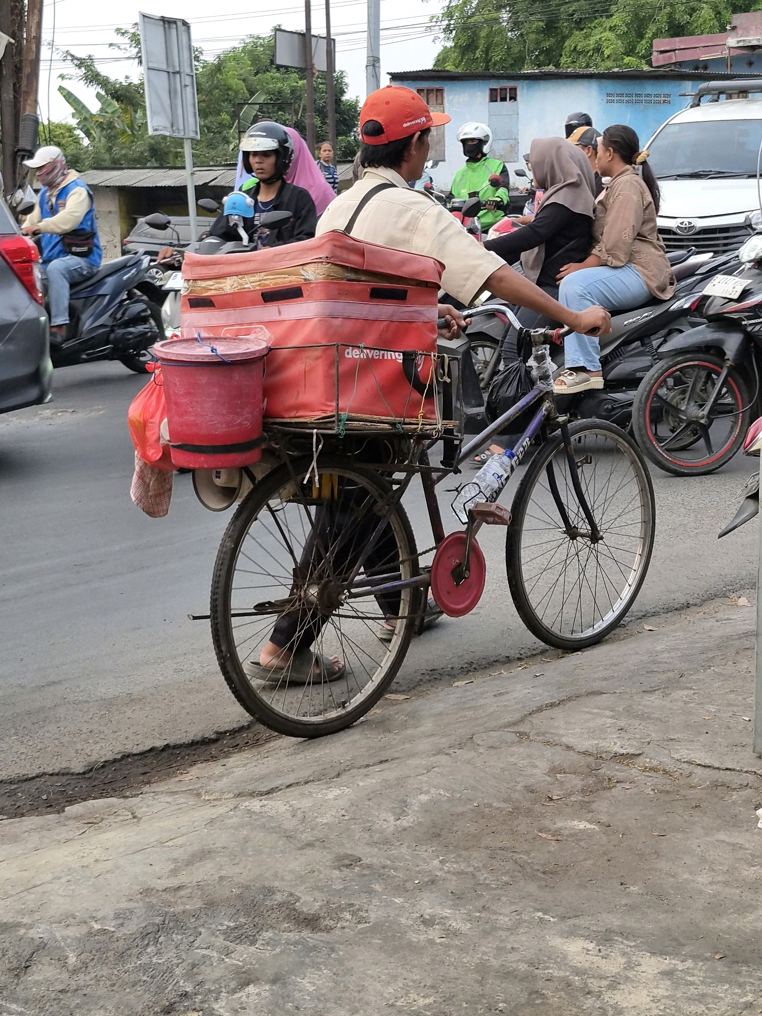 In a street scene in Bekasi, Indonesia, a person wearing an orange cap and a light-colored shirt is riding a bicycle laden with red delivery containers and a red bucket. The bicycle has a bottle of water attached to the frame. The rider is looking ahead and appears to be in motion, navigating through traffic. Several motorcycles are visible around the bicycle, some carrying passengers. A white car is parked in the background. The environment appears to be an urban setting with buildings and trees. The road surface is paved with asphalt and shows some signs of wear and tear. The weather seems to be overcast.