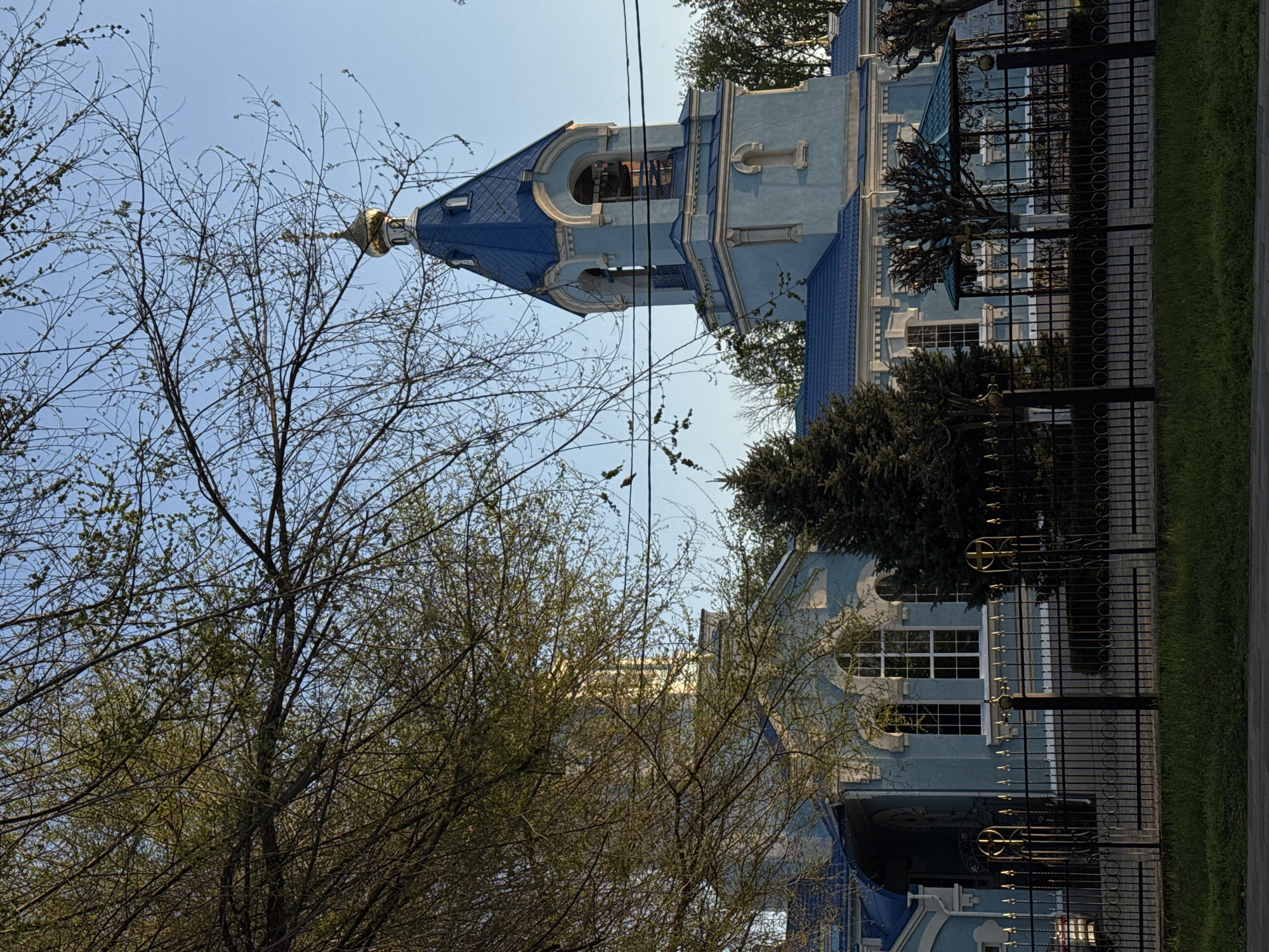 Here is a detailed description of the image:
The image shows a light blue building with a dark blue roof and a golden dome, likely a church or religious building. It is partially obscured by trees with sparse foliage, indicating early spring or late autumn. A black metal fence runs along the side of the building, separating it from the adjacent street or sidewalk. The sky is clear and blue. The image is taken at an unusual angle, rotated 90 degrees, making it appear as if the building is sideways. The location is likely in a city but the specific city cannot be determined from the image.