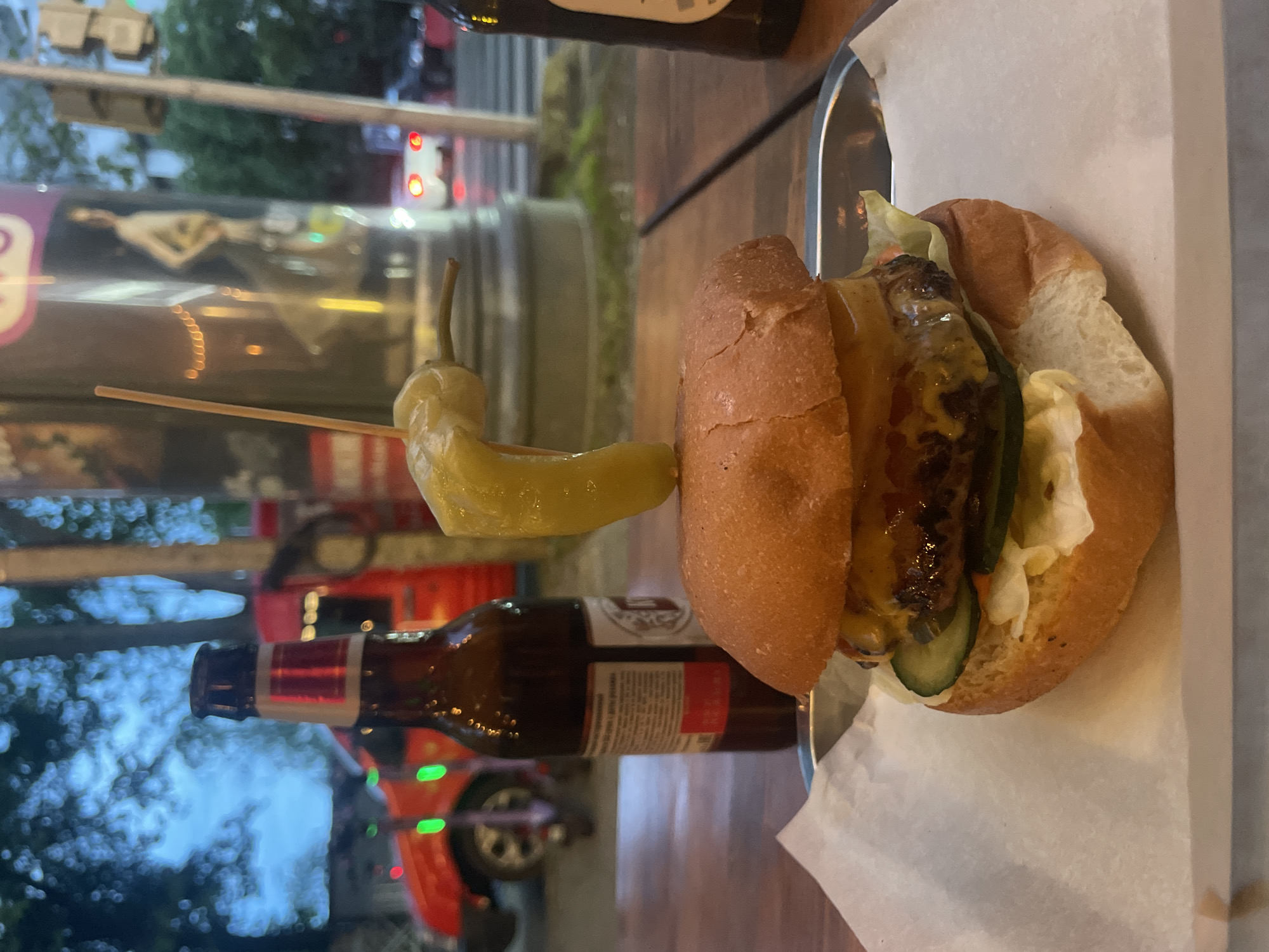 The image shows a hamburger on a wooden table, likely at a restaurant or outdoor dining area. The burger is served on a metal tray lined with white paper. A pickled pepper is attached to the burger with a toothpick. A bottle of beer is also on the table, positioned near the burger. The scene is located in the city of Sochi, Russia, indicated by the Cyrillic script on a sign visible in the background through the glass window. The background reflects an urban setting with streetlights and a glimpse of a red car.