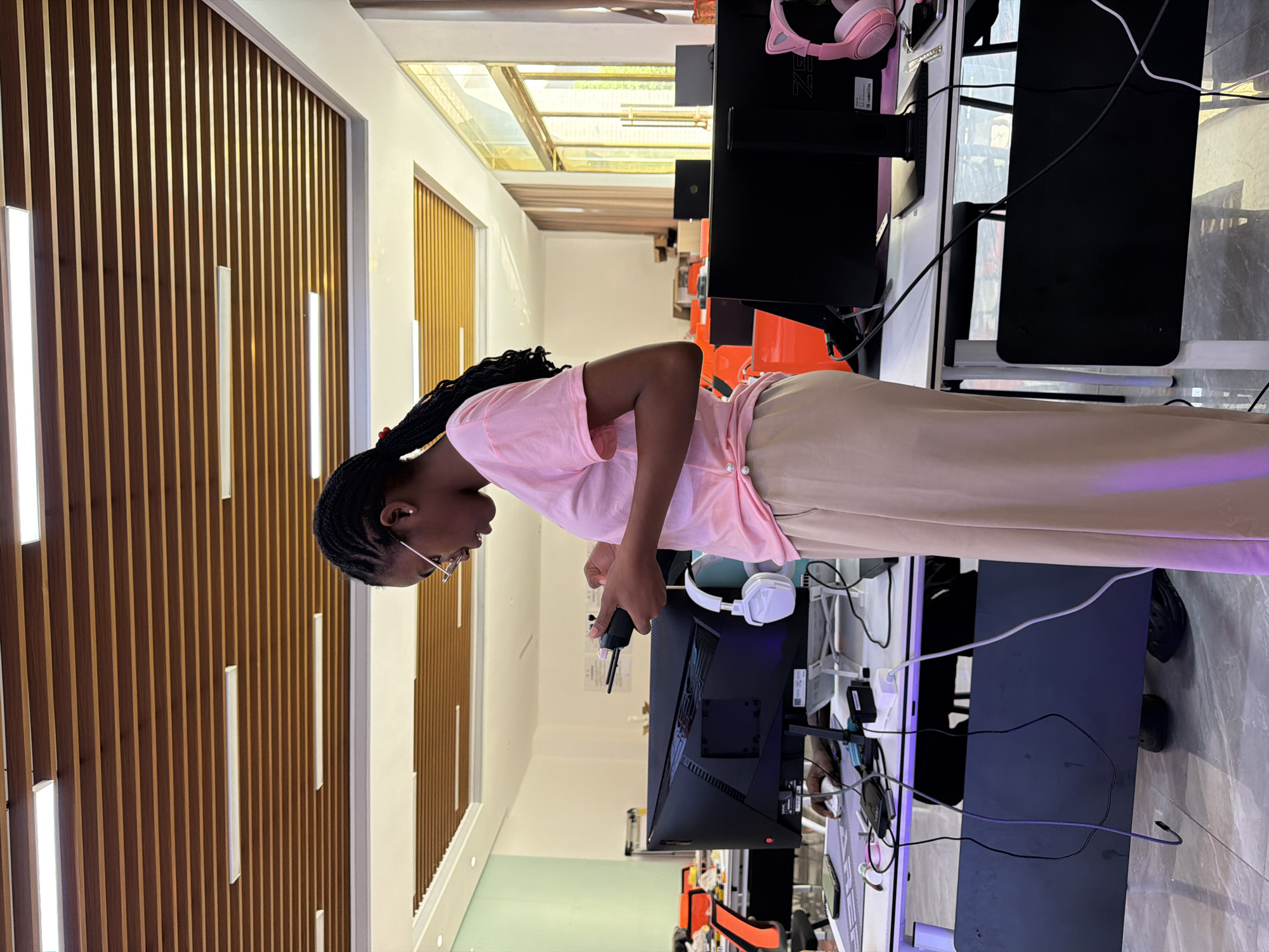 A young Black woman wearing glasses and a pink t-shirt is standing in a brightly lit office or classroom, holding a black remote control in her hands. She is looking down at the remote and appears to be interacting with it. Her hair is braided and falls over her shoulder. She is wearing beige pants. In the background, there are multiple computer monitors and desks, suggesting a workspace or educational setting. The ceiling features a modern design with wooden slats and recessed lighting. The room is spacious and appears to be well-equipped with technology. There are also windows with a view of the outdoors. This scene is located in the city of Jiangjin, China.