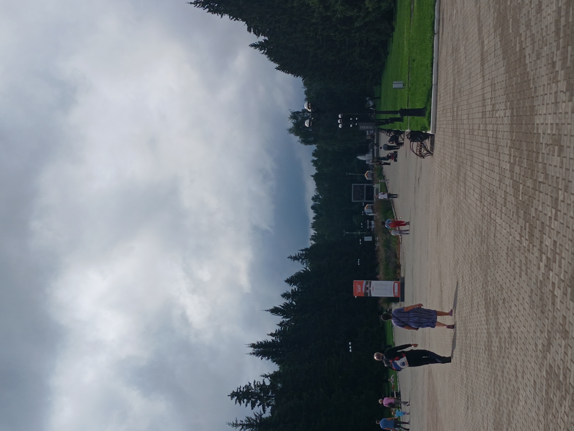 The image depicts an outdoor paved walkway in Yekaterinburg, Russia, with dense evergreen trees lining one side of the path and a grassy area on the other. The sky is overcast with dramatic, dark clouds. Several individuals are walking on the paved area. In the foreground, a man and a woman are walking away from the camera. The man has a backpack, and the woman is wearing a striped dress. Further down the path, more people are visible, some walking and others standing. A tall, vertical sign with text in Russian is present on the left side of the walkway. Streetlights are positioned along the path. In the background, a fountain can be seen, and a Ferris wheel-like structure is partially visible. The overall impression is of a public park or outdoor recreational area on a cloudy day.