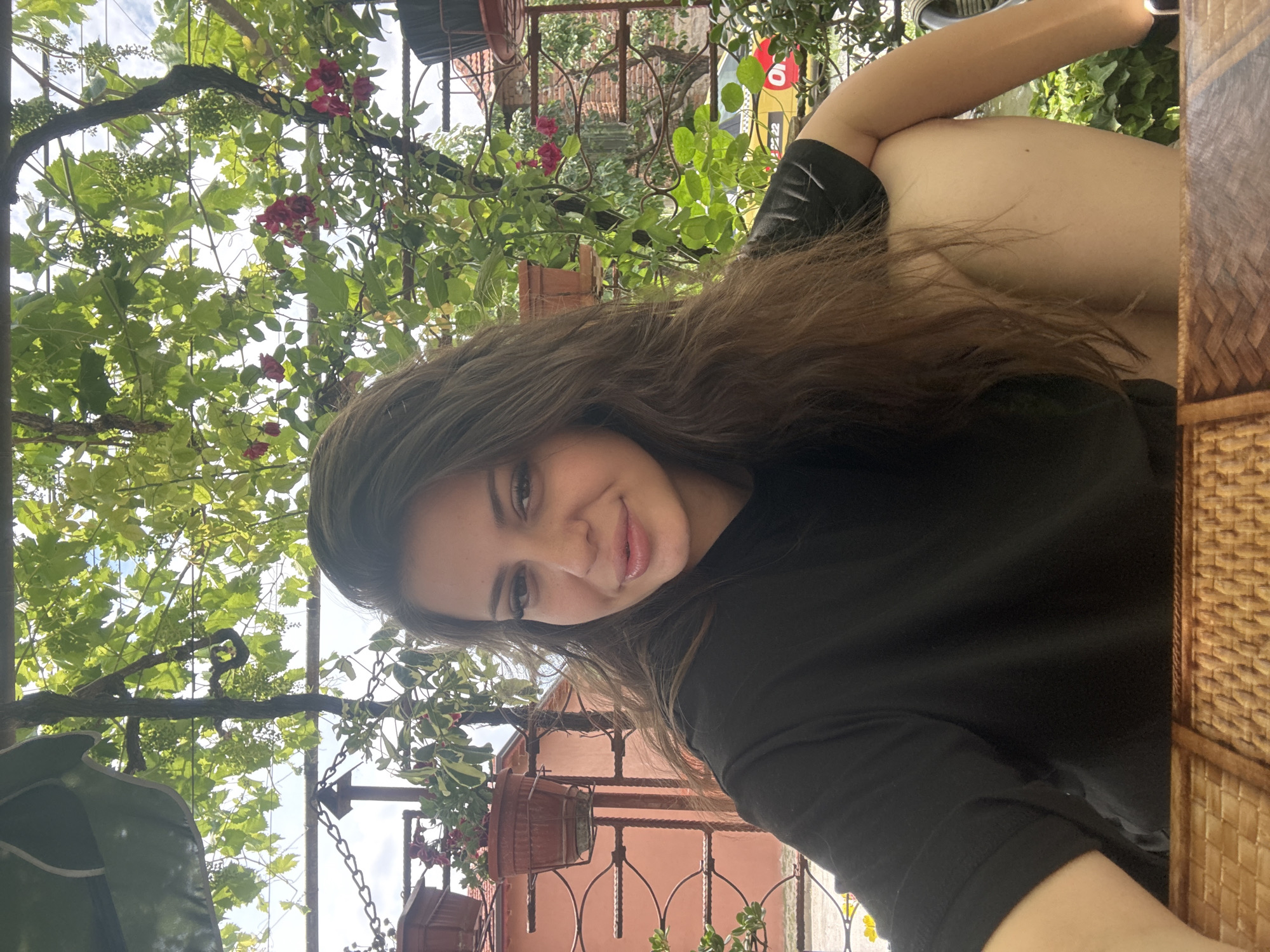 A young woman with long, brown, wavy hair is captured in a medium shot, seated at a table, outdoors. She is wearing a black t-shirt. The background features a lush green canopy of leaves, likely from a grapevine, with some pink flowers visible. Beyond the foliage, a red building with a chain-link fence and potted plants can be seen. The setting appears to be a relaxed outdoor space, possibly a courtyard or patio. The image may have been taken in Govedare, Bulgaria.