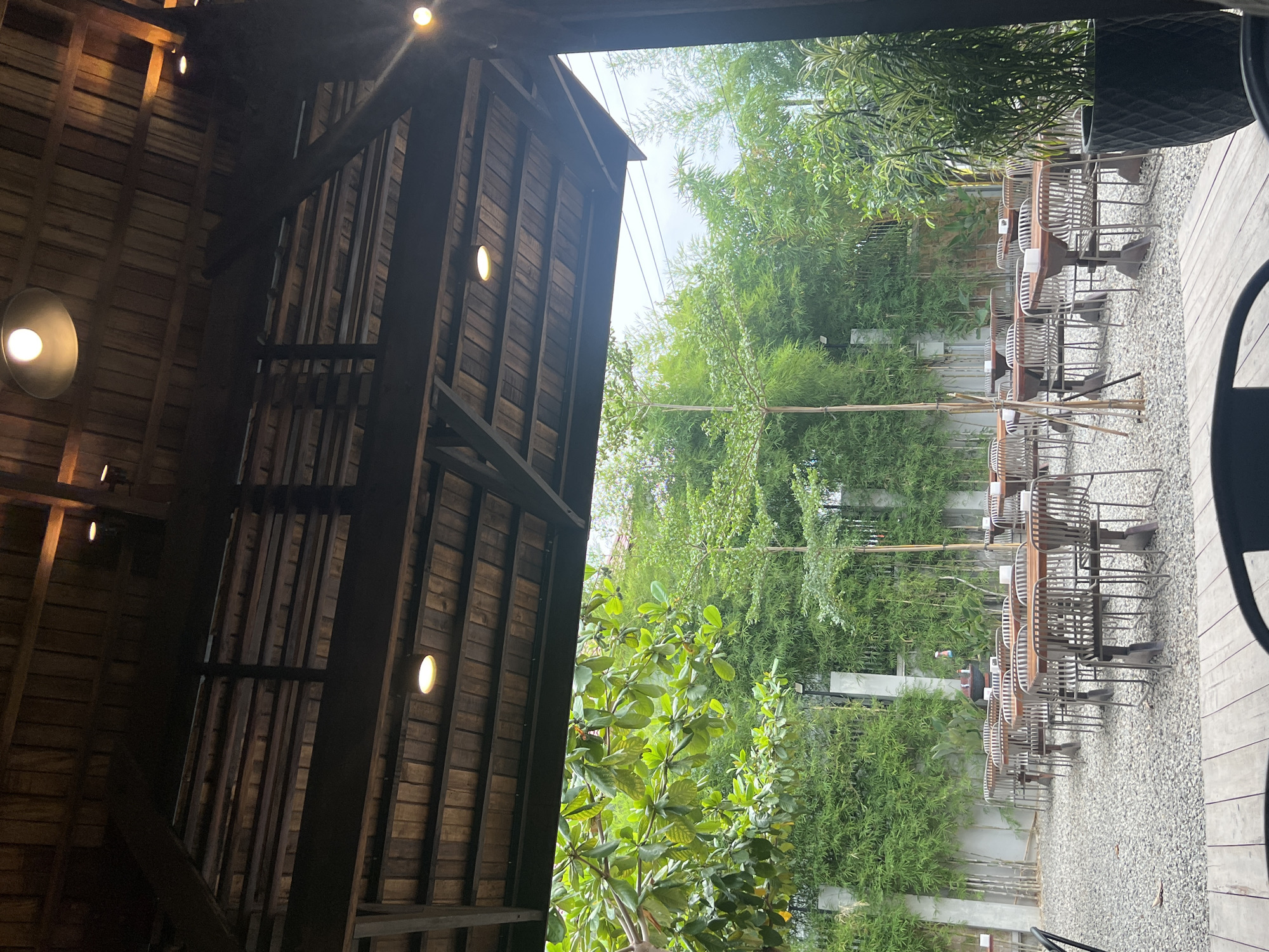 The image depicts an outdoor seating area of a restaurant or cafe located in Bireuenmeunasahdayah, Indonesia. The scene is characterized by a rustic, natural aesthetic.
Dominating the foreground is a wooden deck, with a portion of a black chair visible at the bottom edge, suggesting the vantage point is from within the establishment. Above, a wooden ceiling structure with exposed beams and multiple circular light fixtures is visible.
The main area is covered by a wooden roof supported by dark, sturdy beams, providing shade. Through the opening of this roof, a lush green outdoor space is revealed.
The ground outside is covered in grey gravel. Rows of tables with wooden tops and metal chairs with intricate metalwork are neatly arranged on the gravel. Some tables have small objects on them, possibly for decoration or service.
A dense wall of bamboo and other green foliage forms a natural backdrop behind the seating area. Several tall, slender trees with sparse foliage are interspersed among the bamboo. Large, smooth grey concrete pillars punctuate the greenery in places. In the mid-ground, a dark planter containing a tall, leafy plant stands to the right. The overall atmosphere is tranquil and inviting, suggesting a relaxed dining or gathering space surrounded by nature.